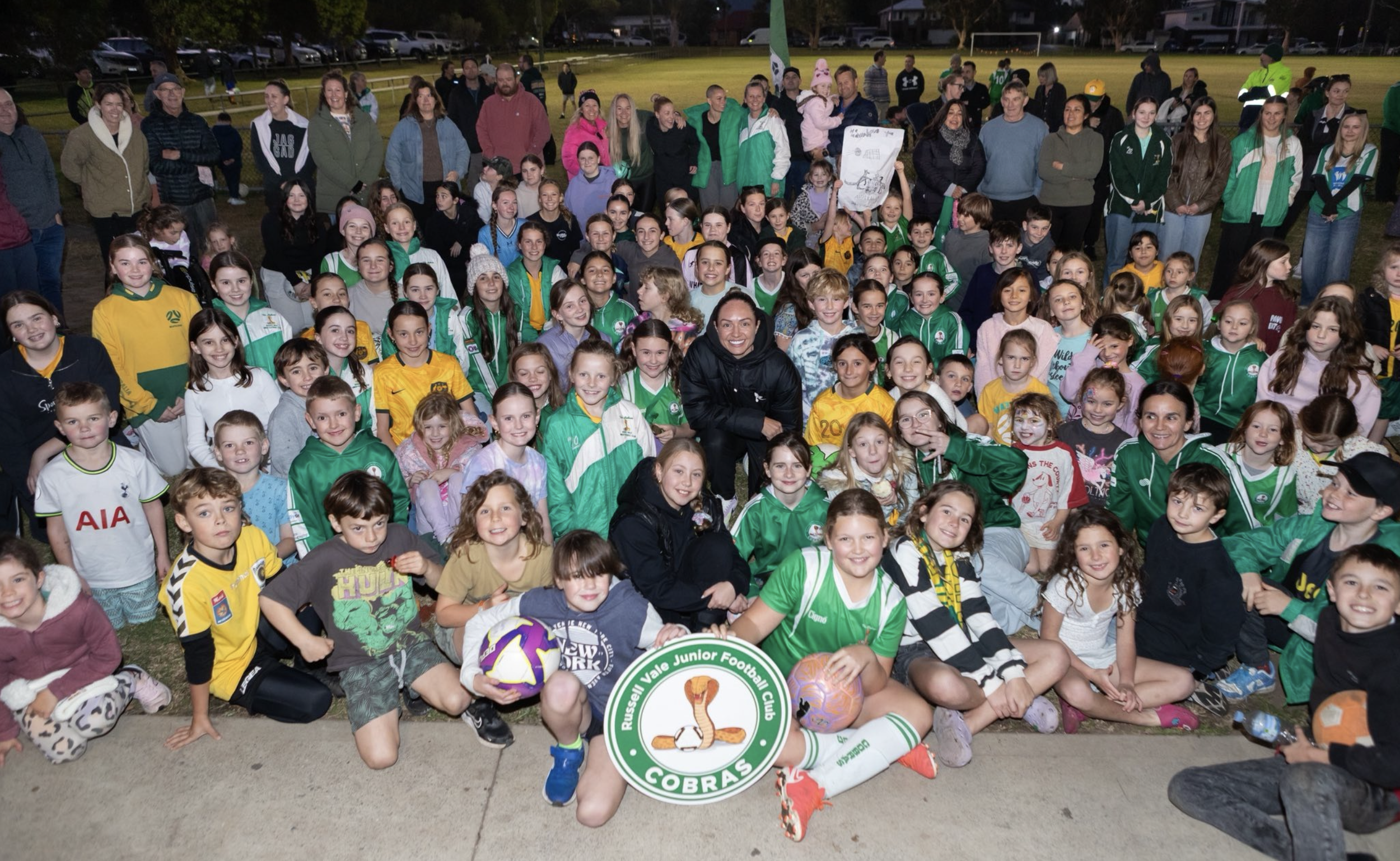 Group photo of children and adults at Russell Vale Junior Football Club, with some holding footballs, outdoors in a park or sports field during the evening.