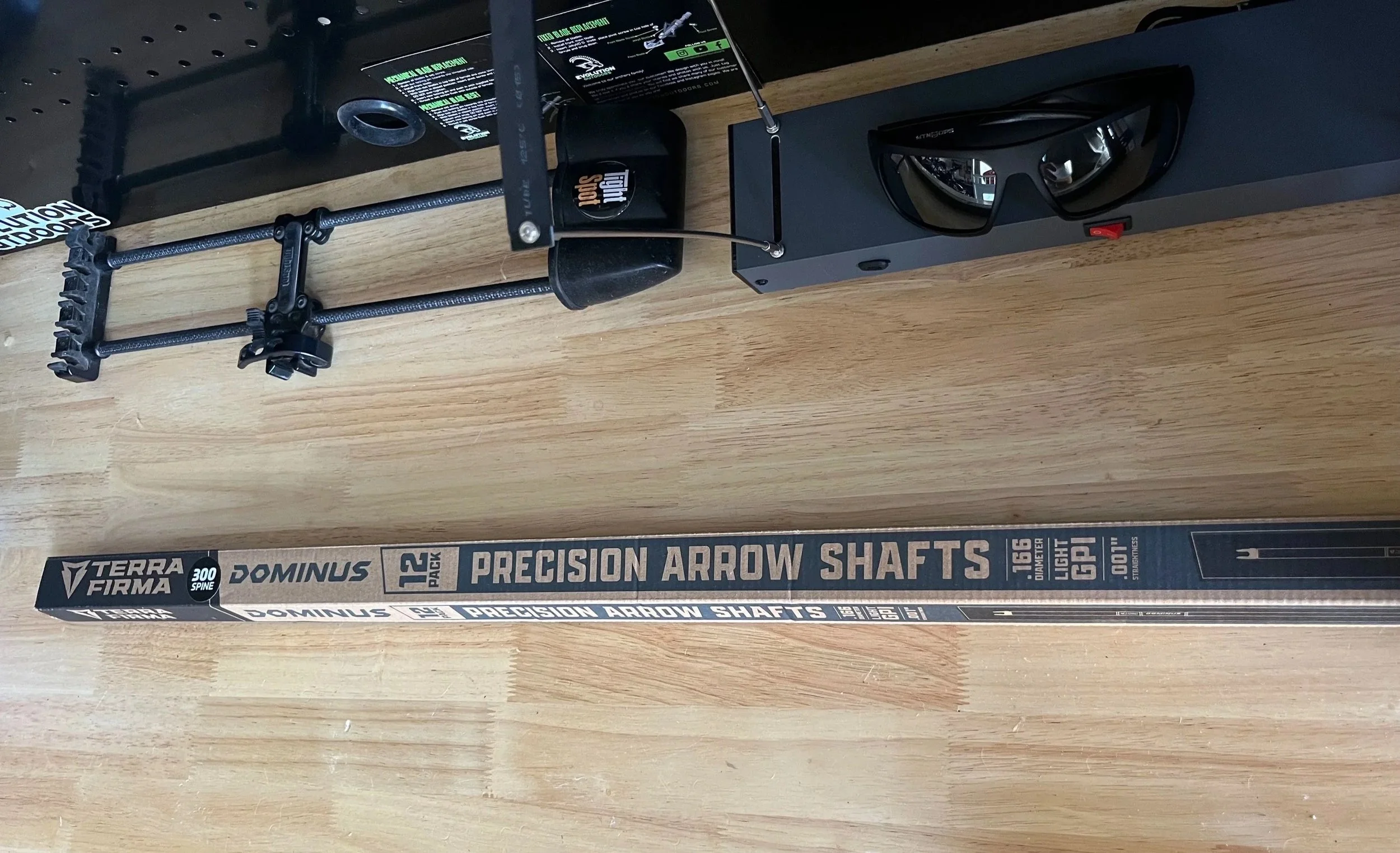 A box of Terra Firma Dominus 12 pack precision arrow shafts, a mechanical bow release, and a pair of black sunglasses on a wooden surface.