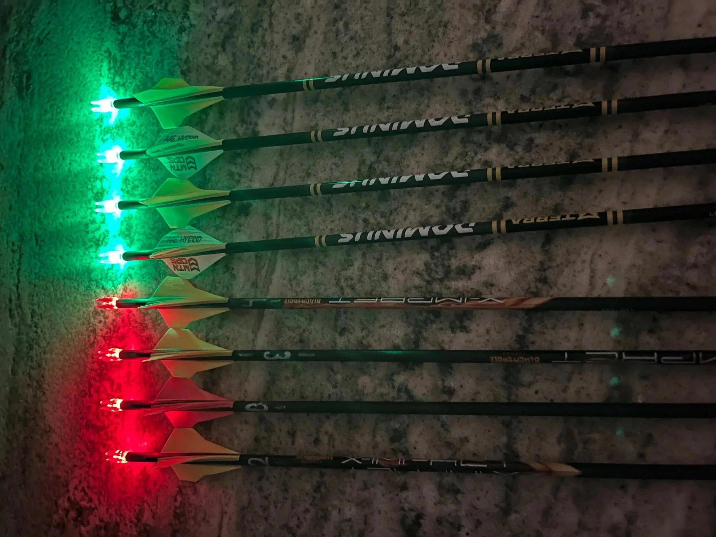 Set of six illuminated custom arrows with lighted nocks, (green and red) on a rough ground surface, arranged in a row.