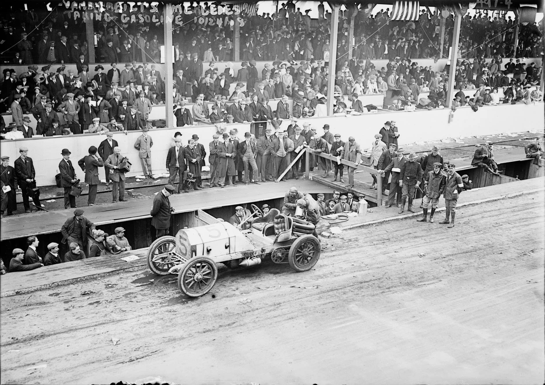 Black and white photo of a crowd of people at a racing event, gathered behind a barrier, with some standing and others sitting. The crowd is mostly men wearing hats and coats, with a few women visible. In the foreground, there is a vintage race car with a large number 5 on its side, on a dirt track. Some individuals are holding cameras and papers, and there are officials or officials on the track.