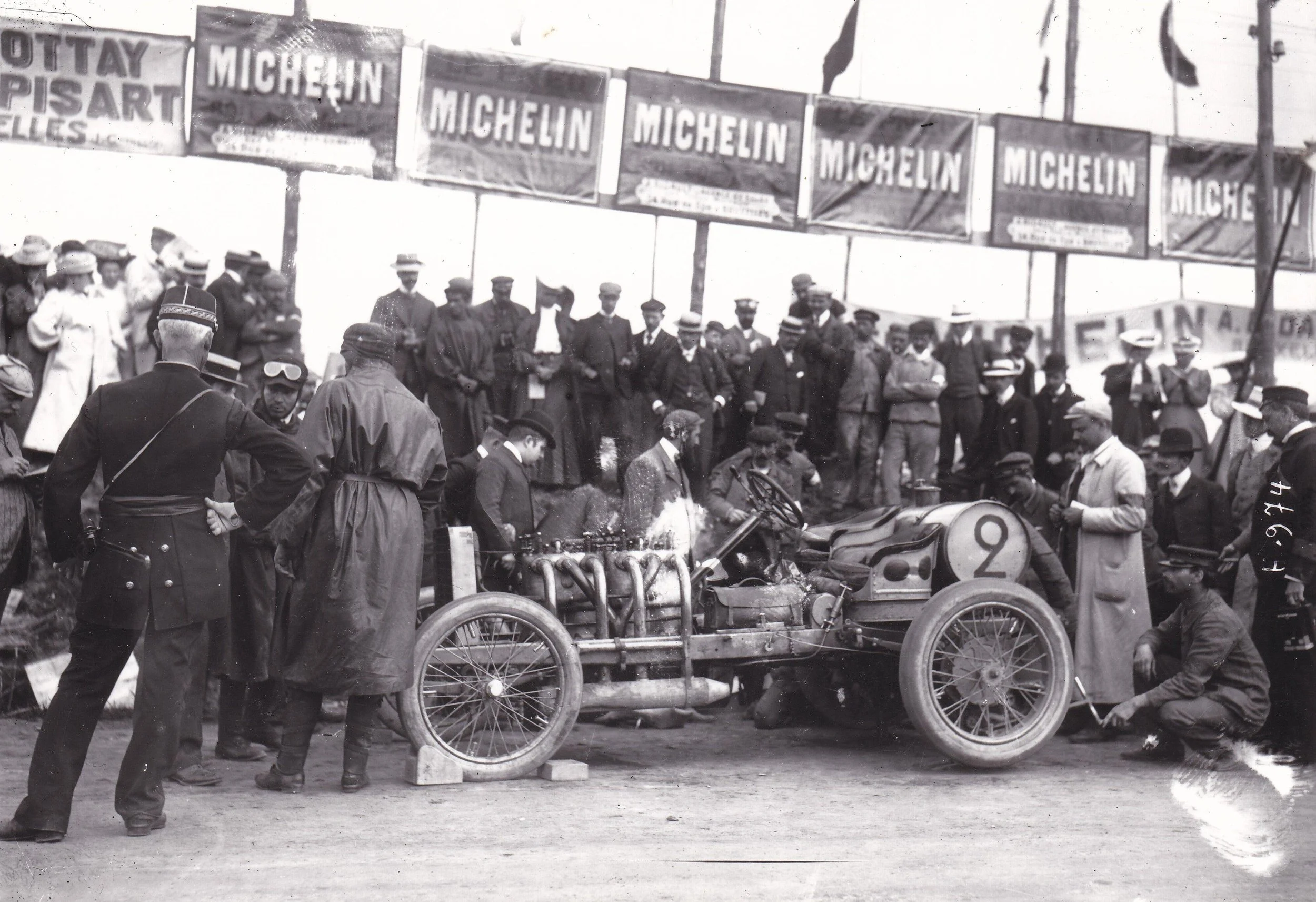 A vintage black-and-white photograph of a crowd attending an event featuring early motor racing. There are several men and women gathered around, some dressed in period clothing. A race car with a visible number 92 is in the foreground, with a mechanic or official attending to it. Large banners overhead advertise Michelin tires, and a few flags are flying in the background.