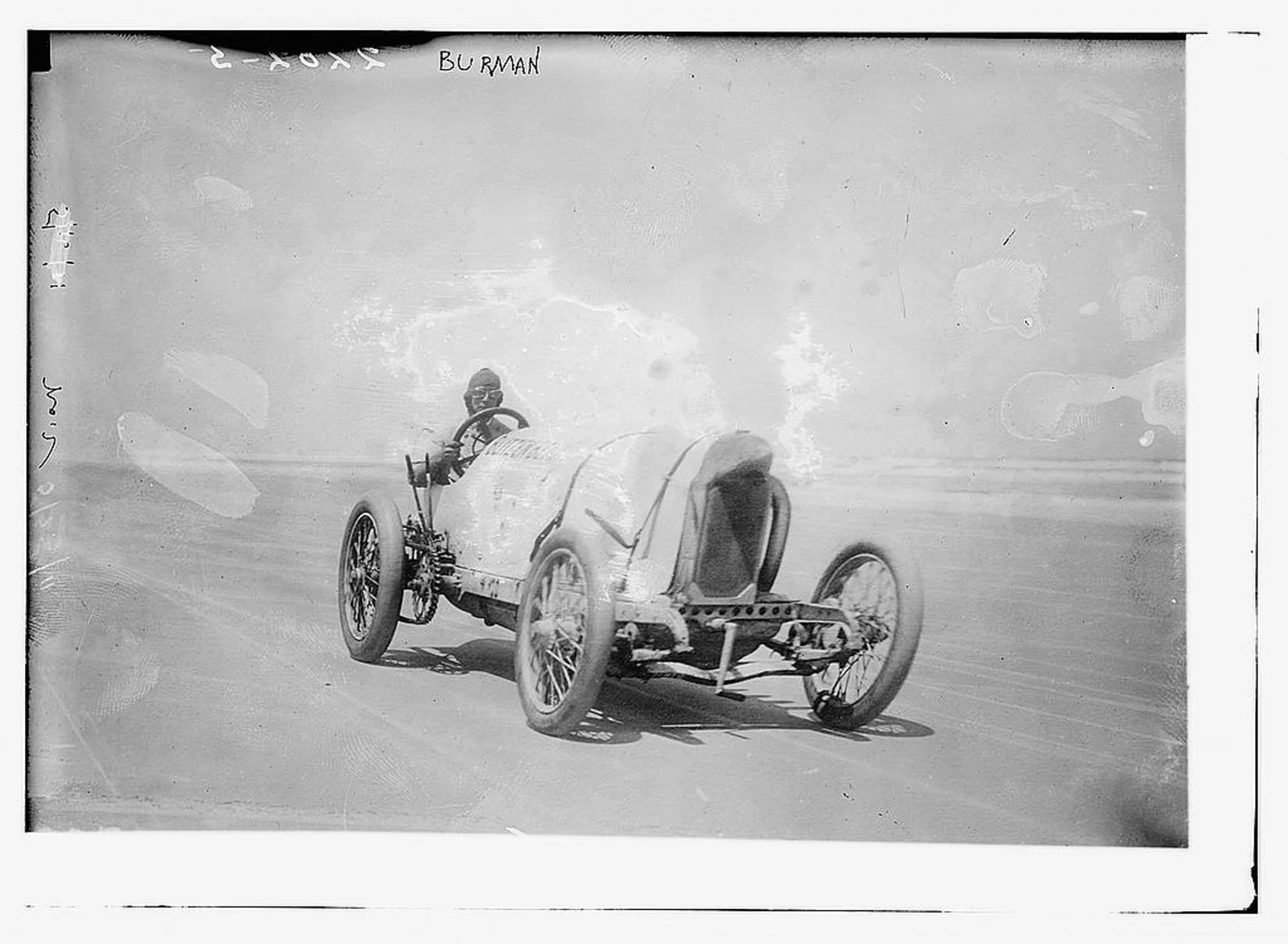 Black and white photograph of a vintage race car with a driver, speeding on a track against a clear sky background.