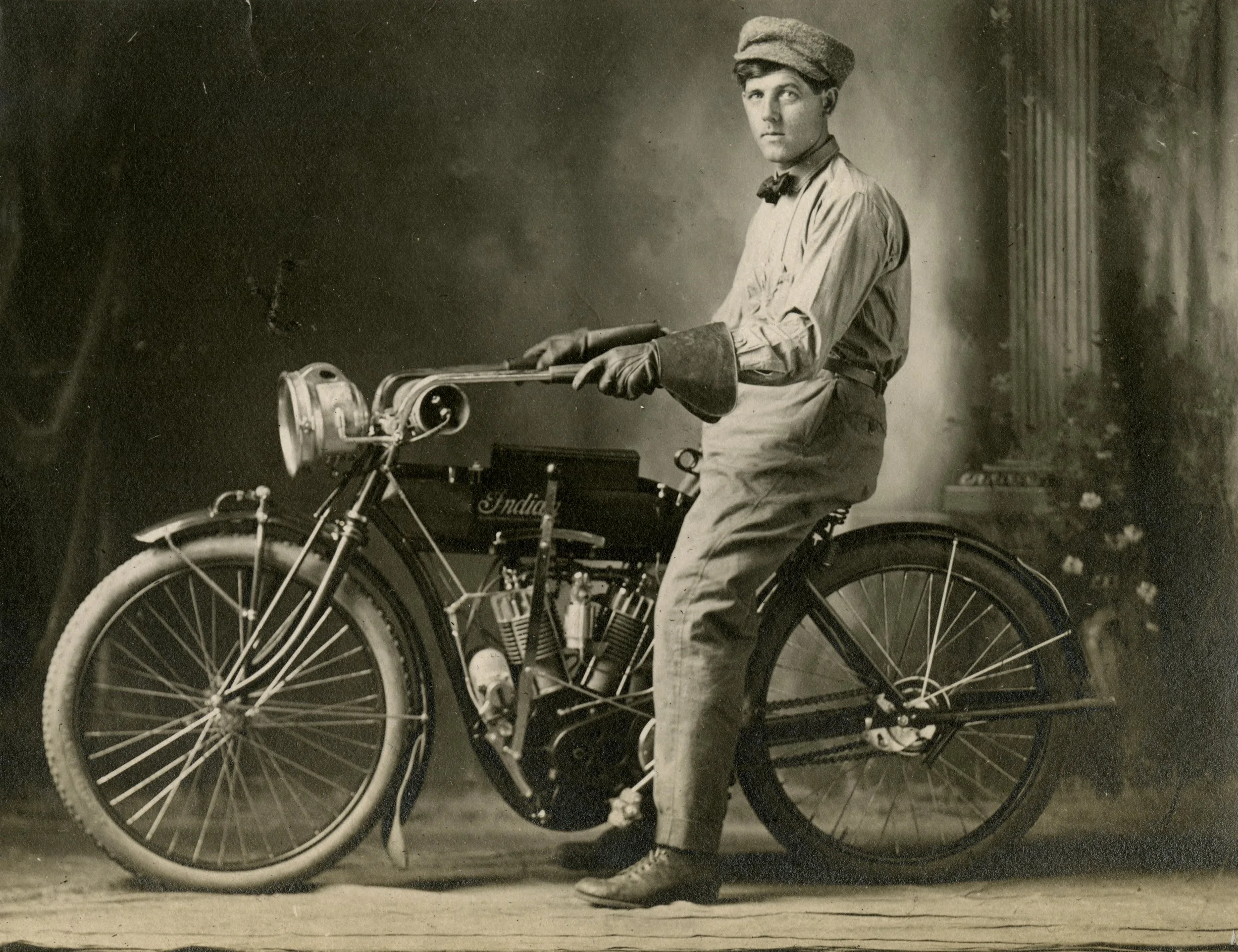 Old black and white photograph of a man in vintage clothing sitting on a motorcycle, holding the handlebars, and looking at the camera.