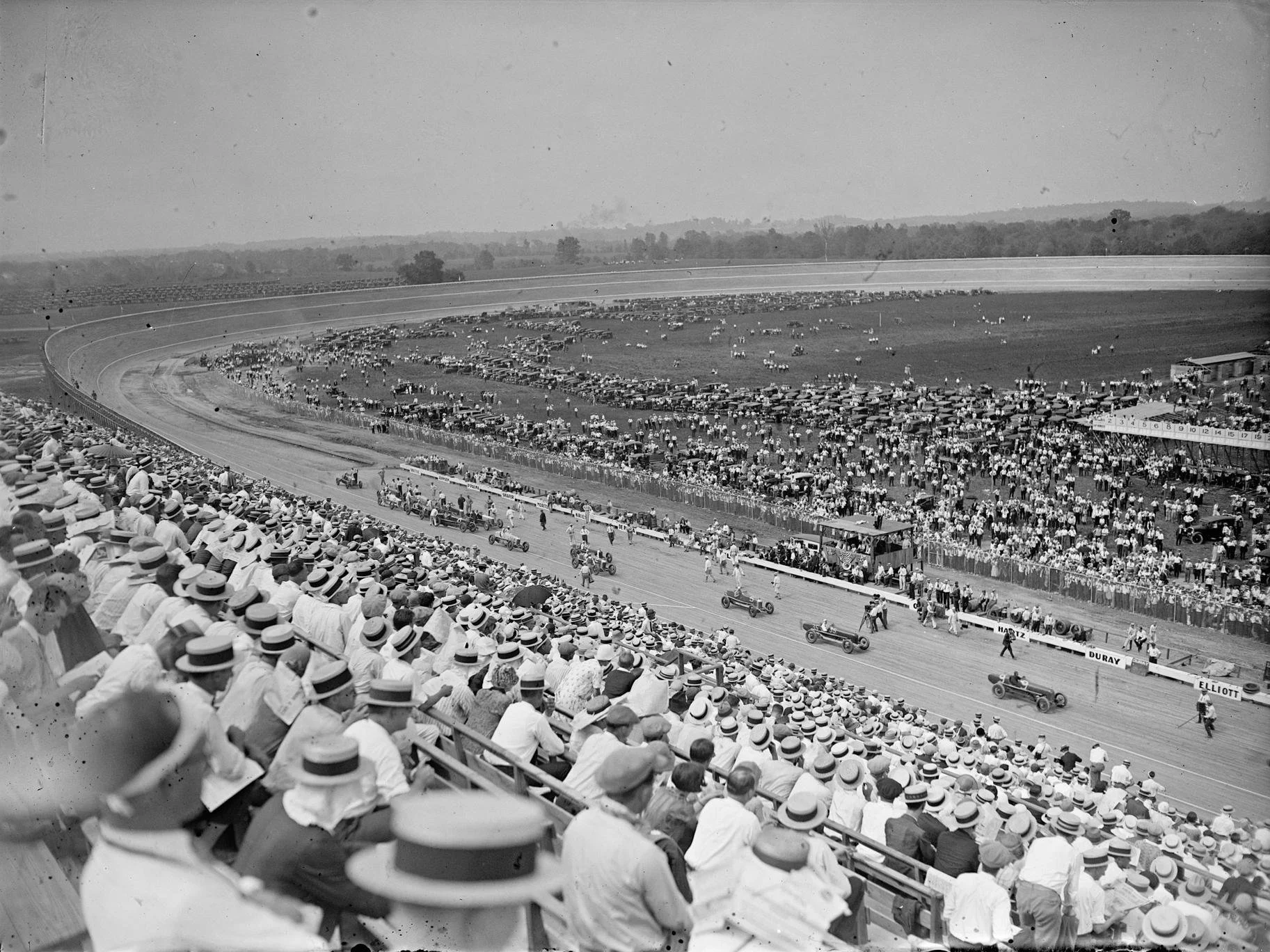Black and white photo of a crowded racetrack with spectators wearing hats watching a race. Cars are on the track and people are on the infield.