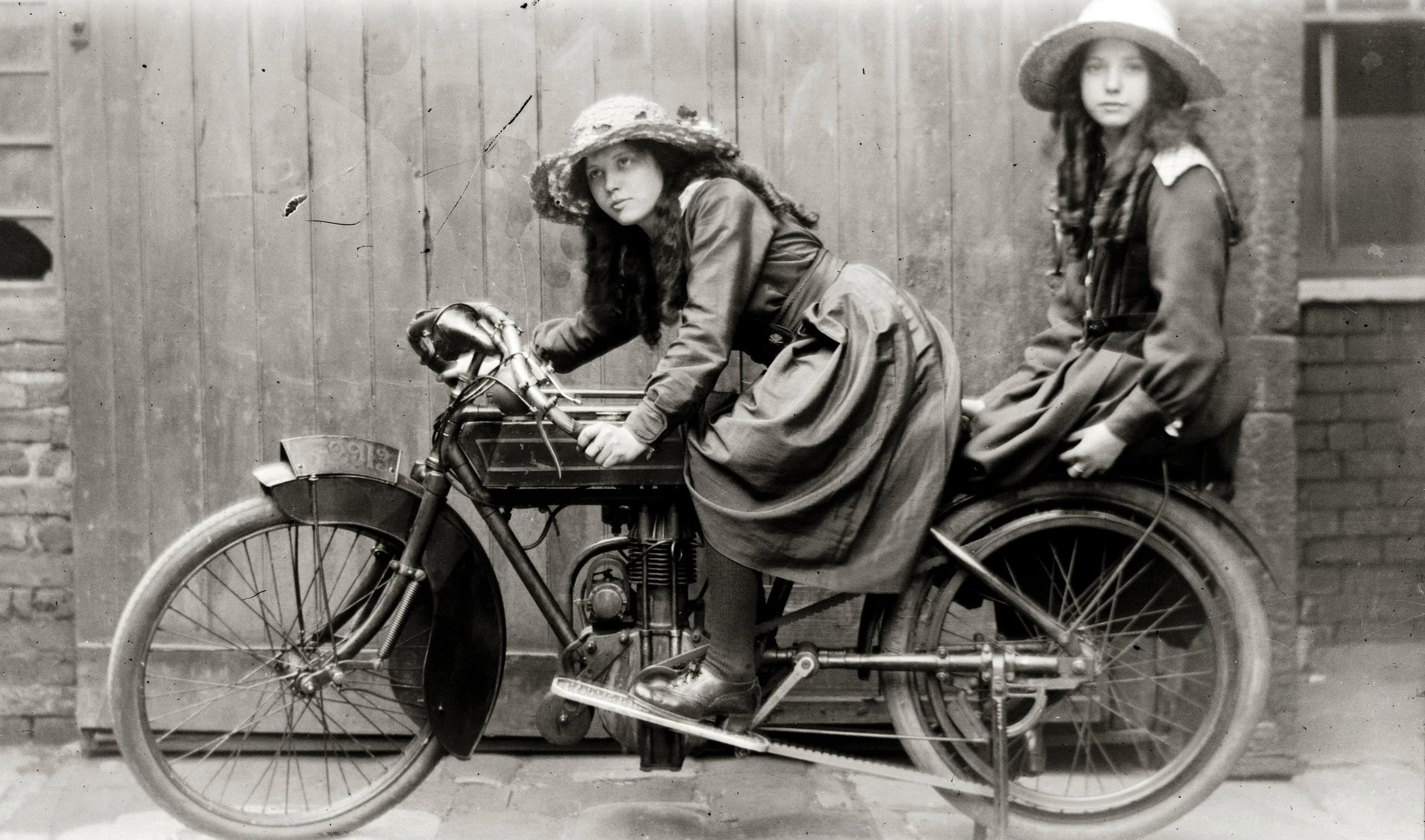 Two young girls with long hair and hats, sitting on a vintage motorcycle, in front of a wooden fence.