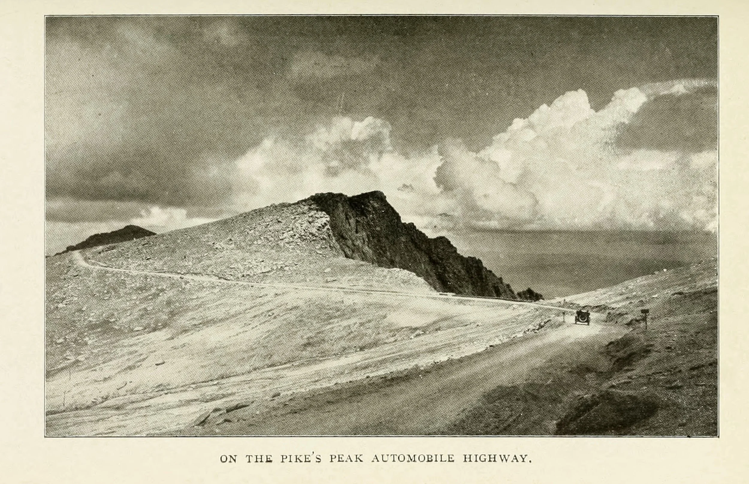 Black and white photograph of a mountain landscape with a winding road and an automobile on the Pike's Peak automobile highway, with a cloudy sky overhead.