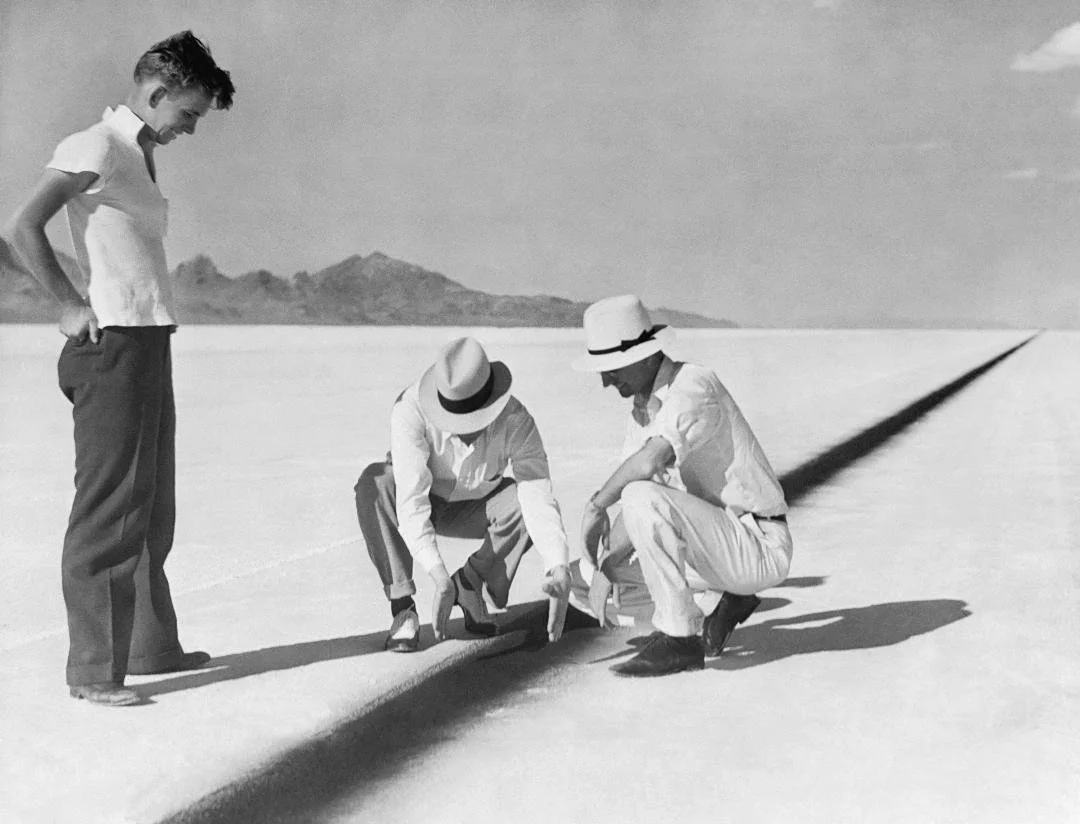 Three men examining a track on a vast salt flat with mountains in the distance, all dressed in light clothing and hats, on a sunny day.