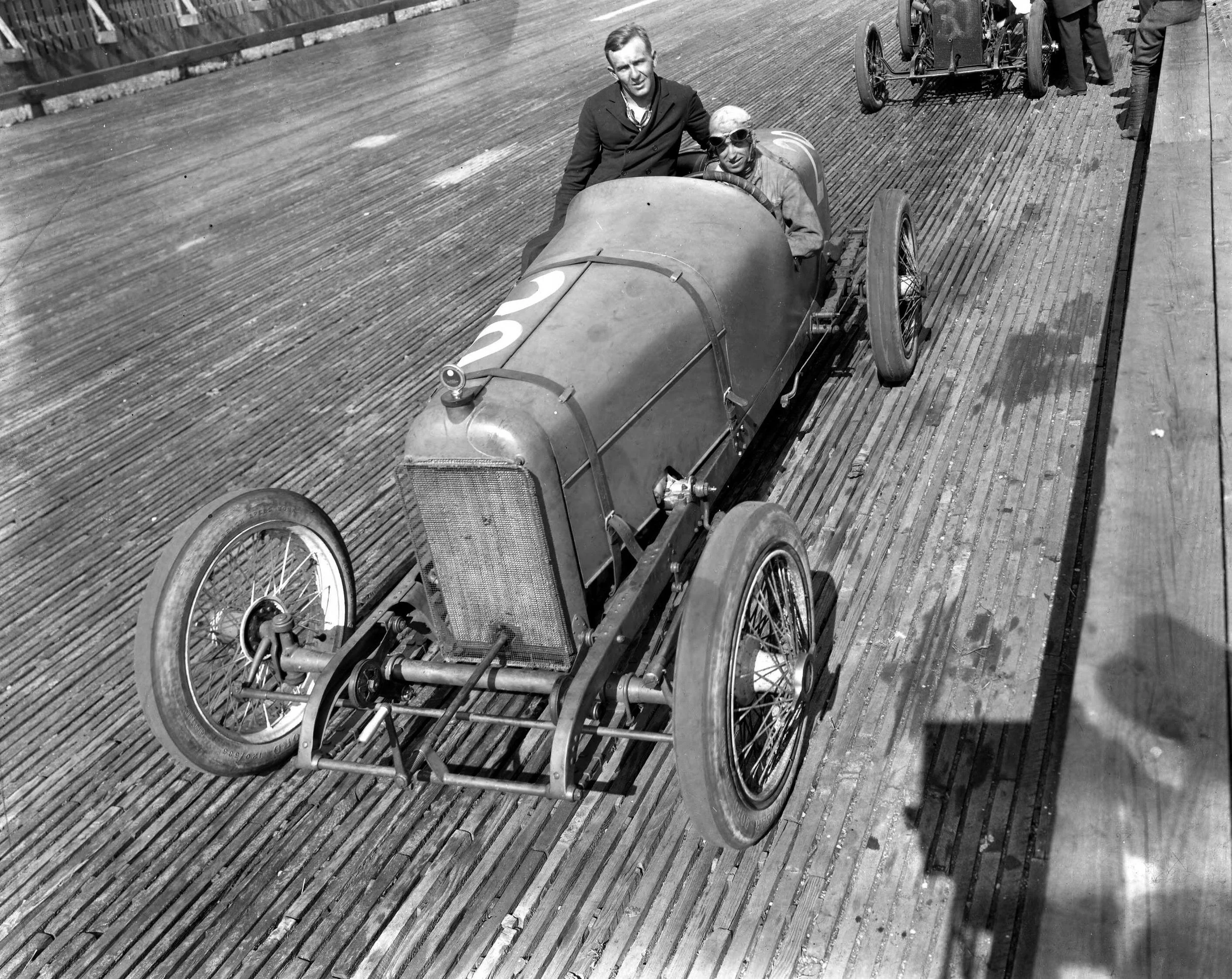Black and white photo of two men next to a vintage race car on a wooden track. One man is sitting in the car wearing goggles, and the other is standing beside the car. There are other race cars and people in the background.