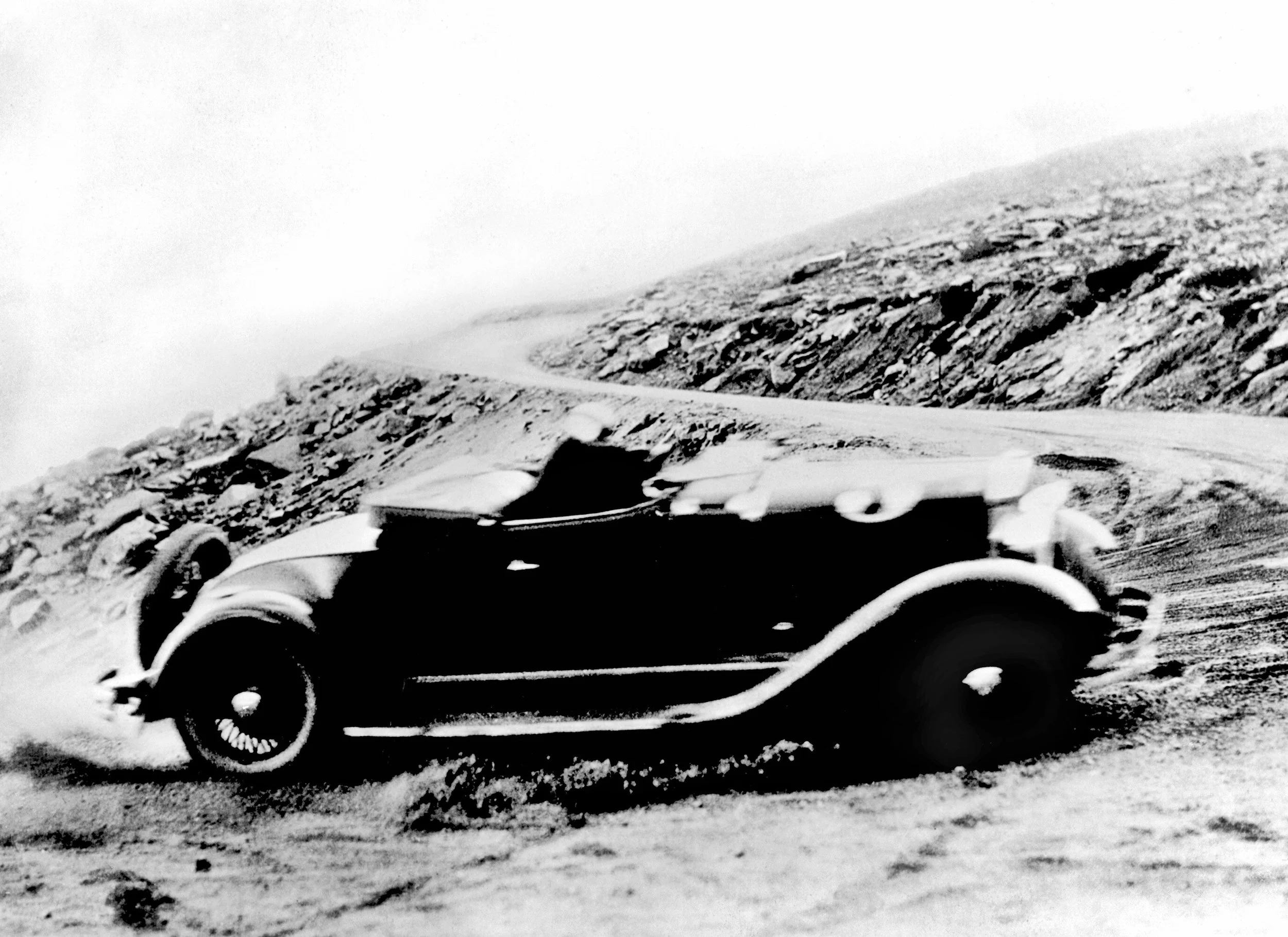 Black and white photo of a vintage car driving up a mountain road track.