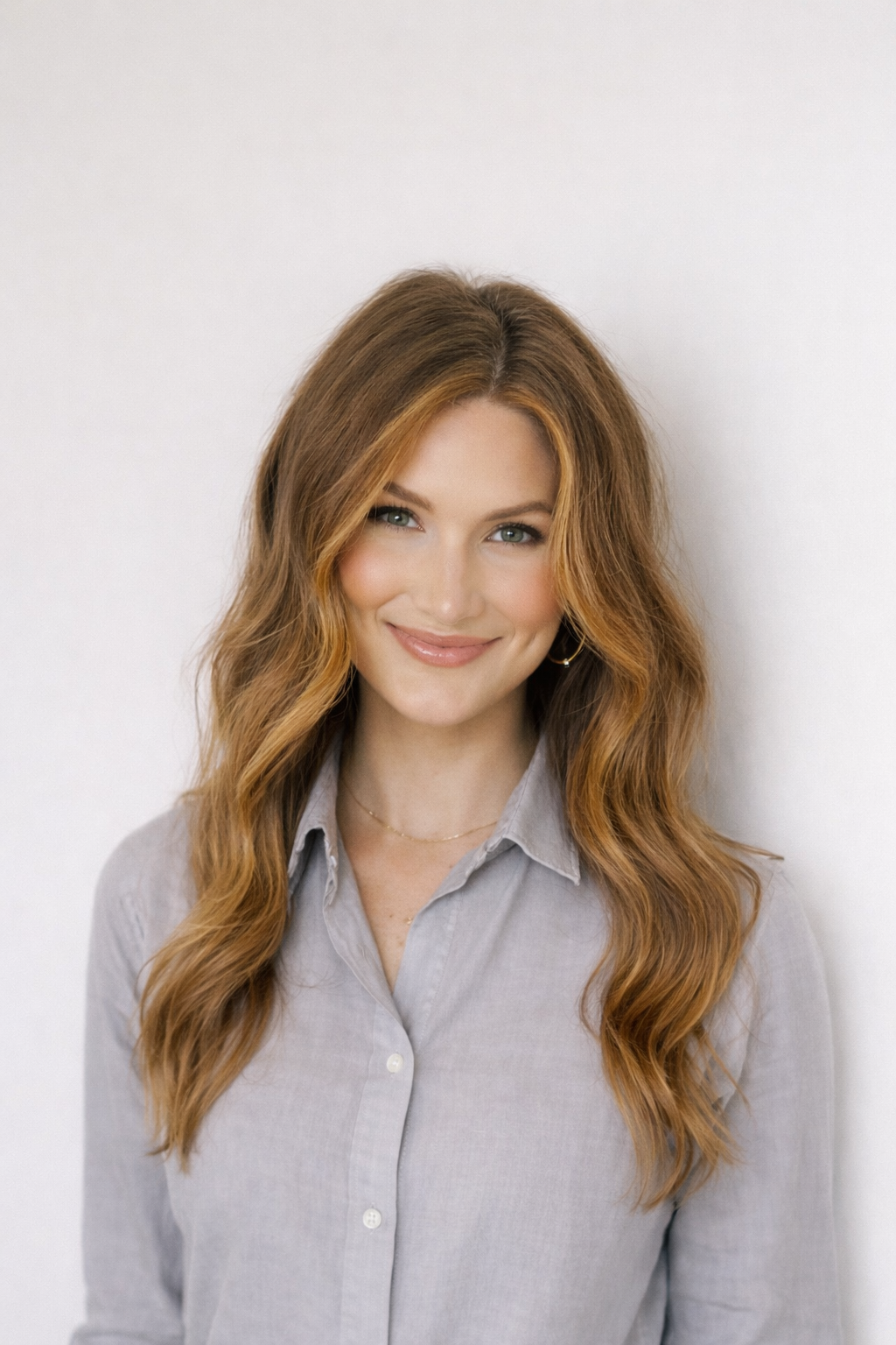 A woman with long, wavy red hair and blue eyes smiling at the camera, wearing a light gray button-up shirt and delicate jewelry, against a plain white background.
