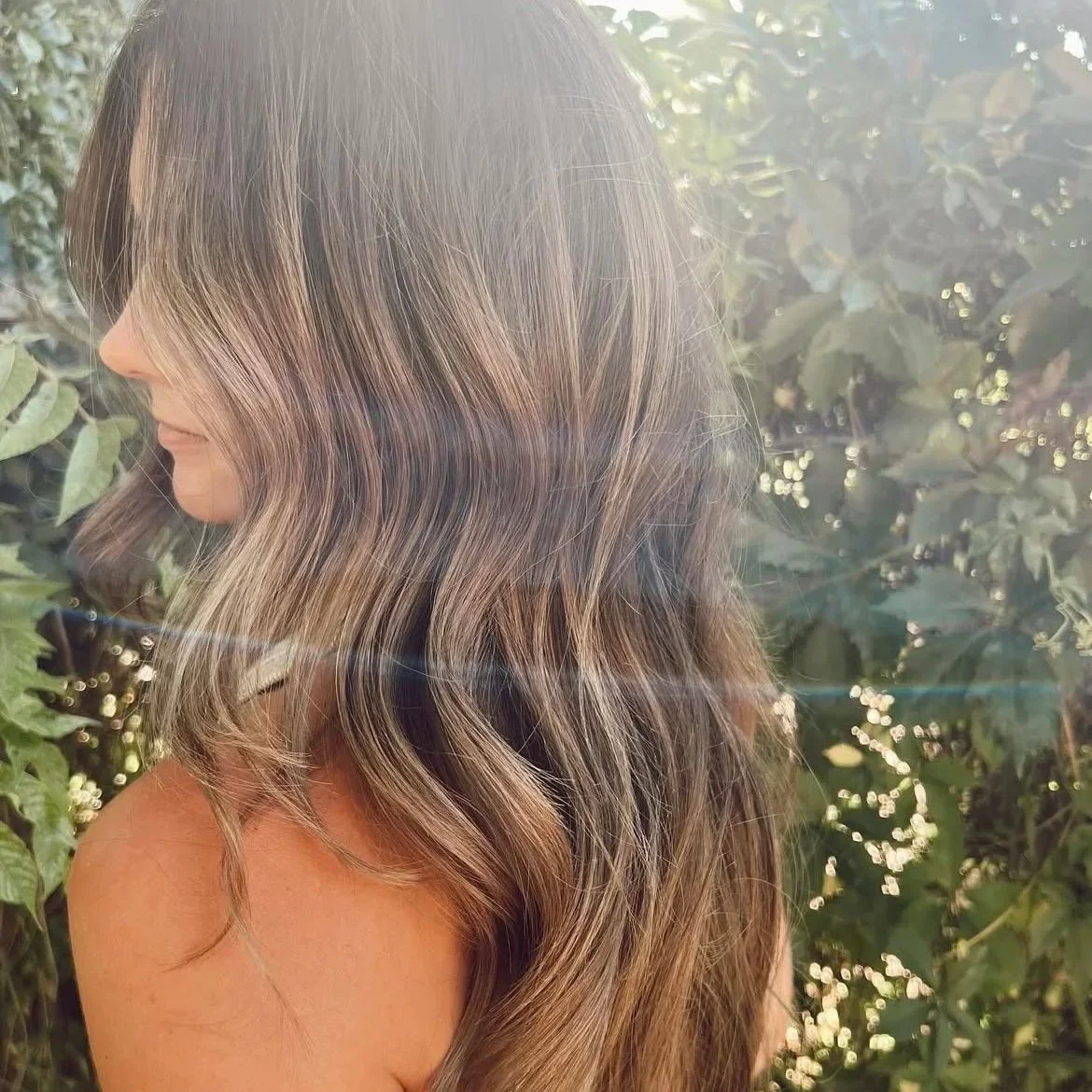 Side profile of a woman with long, wavy brown hair with blonde highlights, standing outdoors among green foliage with sunlight shining through.
