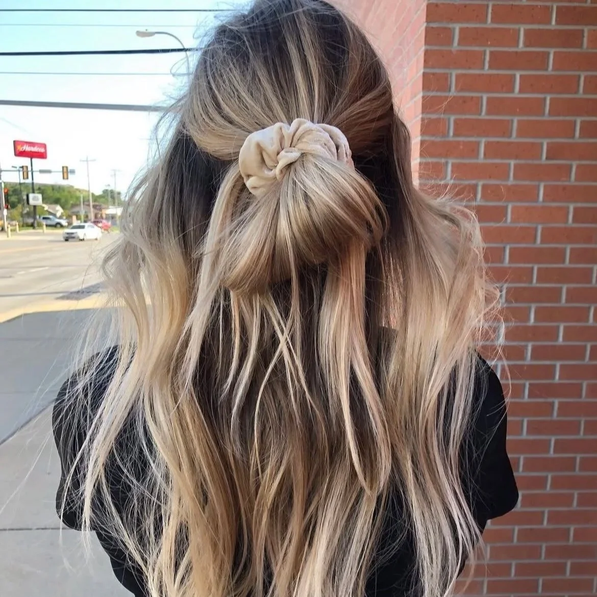 Woman with long, wavy blonde hair tied back with a beige scrunchie, standing outside near a brick wall.