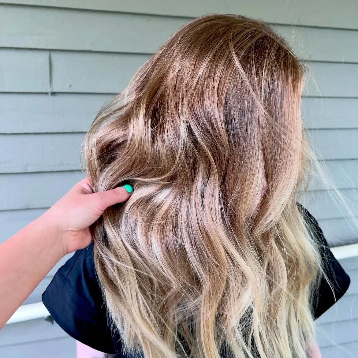A person with long, wavy, blonde hair with subtle pink highlights, holding a section of it.