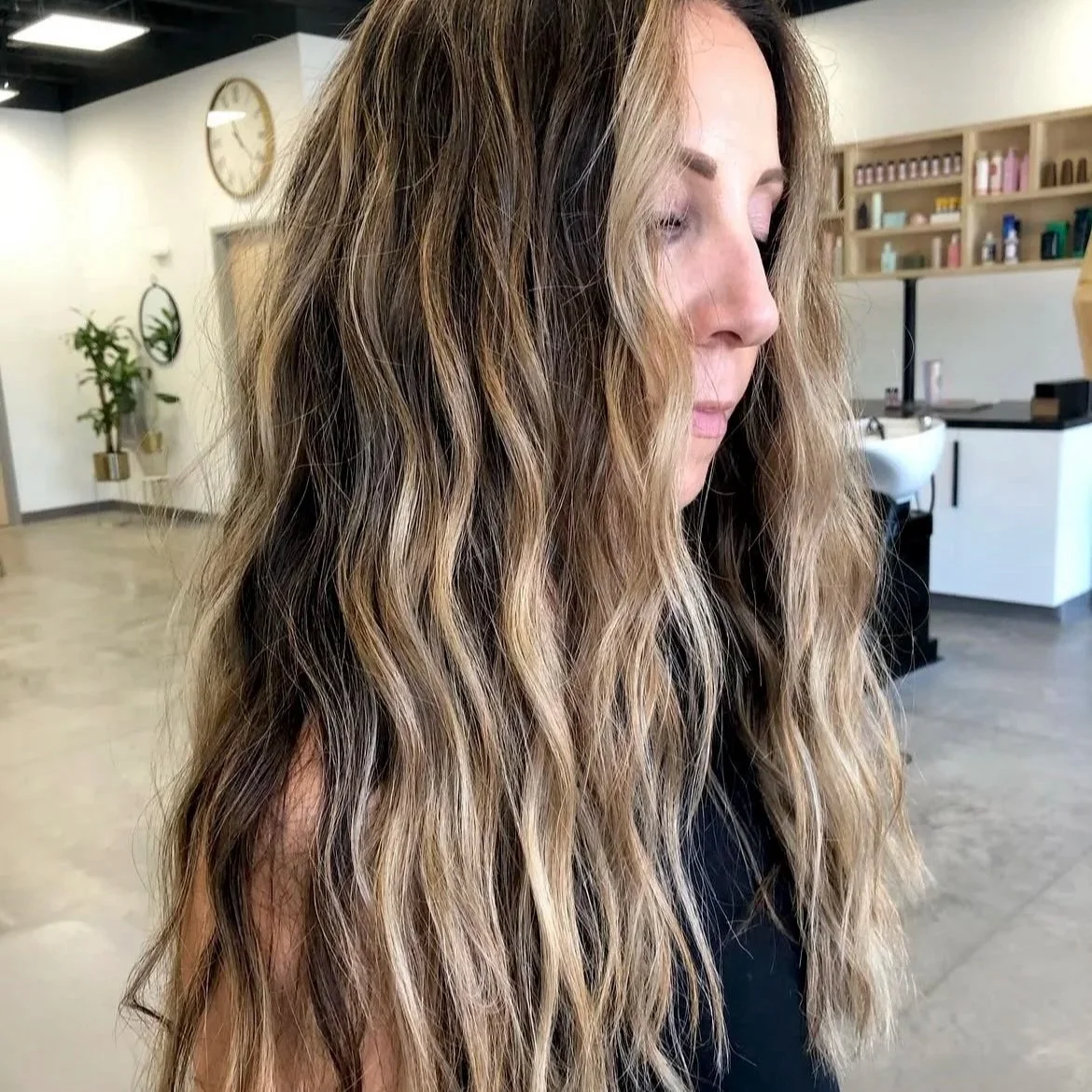A woman with long, wavy, blonde hair with darker roots, standing in a modern salon with plants and shelving in the background.