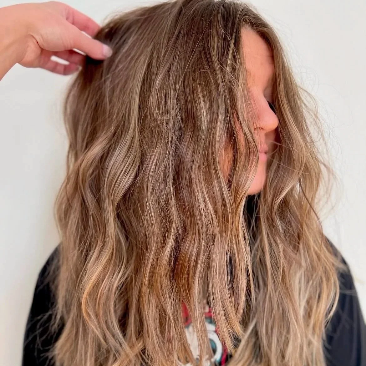 A woman with long, wavy, light brown hair being styled or adjusted by a hand.