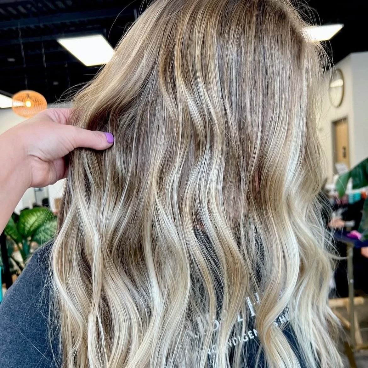 Blonde hair with loose waves held by a hand, in a salon or indoor setting.