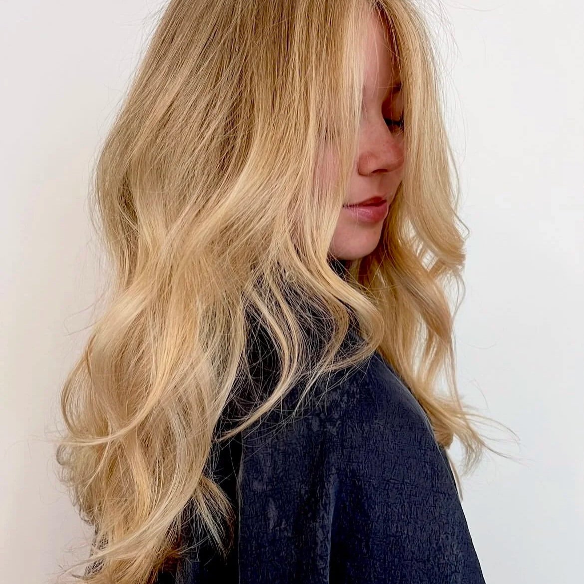 A woman with long, wavy blonde hair looking down, wearing a dark jacket against a plain white wall.