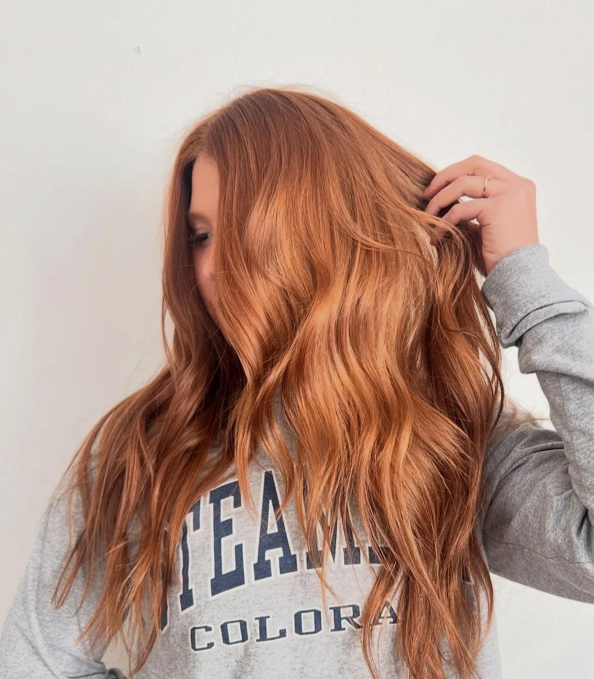 A woman with long, wavy red hair, wearing a gray sweatshirt with text, touching her hair with her right hand, and standing against a plain white background.