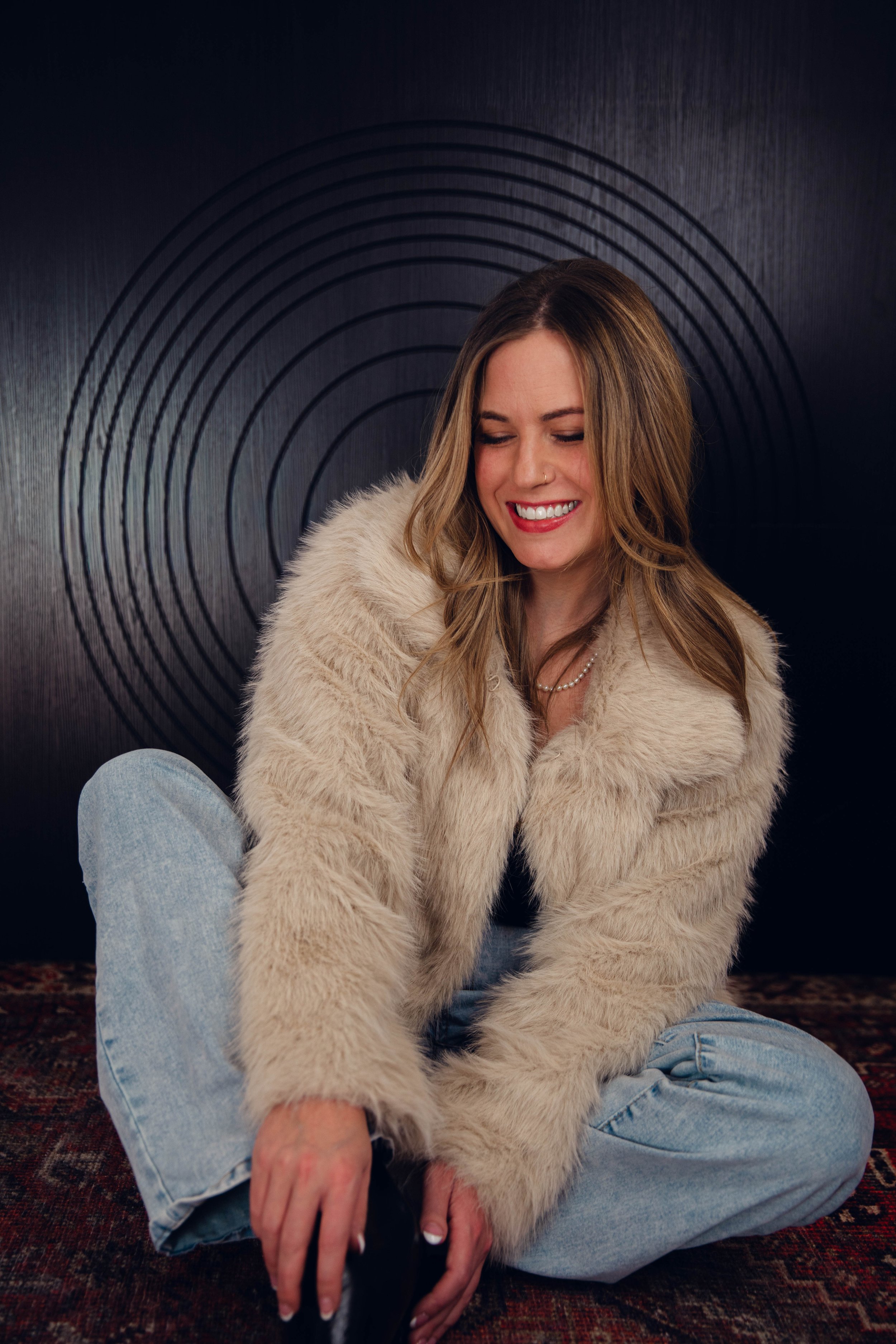 A woman with long wavy hair, wearing a beige faux fur coat, light blue jeans, and a pearl necklace, sitting cross-legged on a patterned rug and smiling with eyes closed against a black wall with concentric circle patterns.