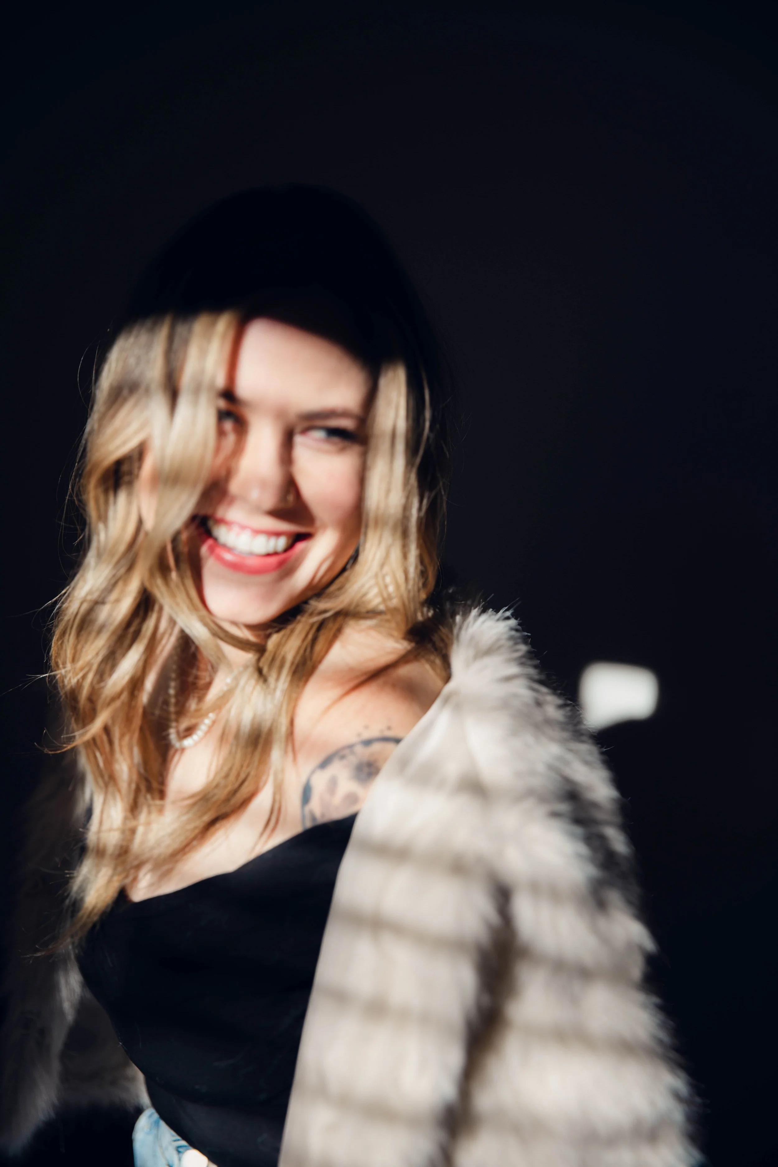 A woman with wavy blonde hair, smiling and turn suggests she is laughing or in a joyful mood, wearing a black top and a light-colored fuzzy coat, against a dark background.