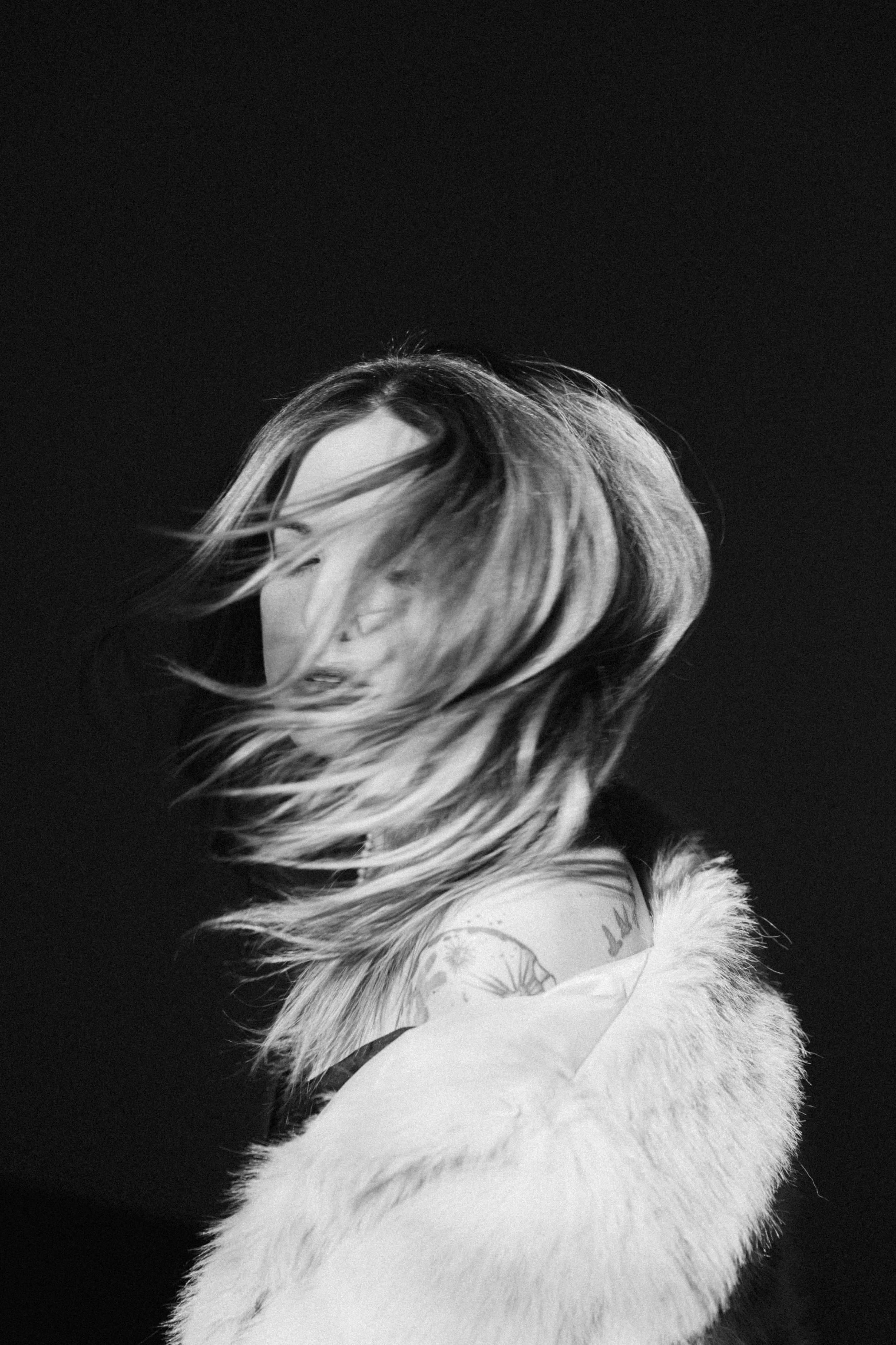 A black and white photo of a woman with medium-length hair blowing in the wind, wearing a fur coat, against a dark background.