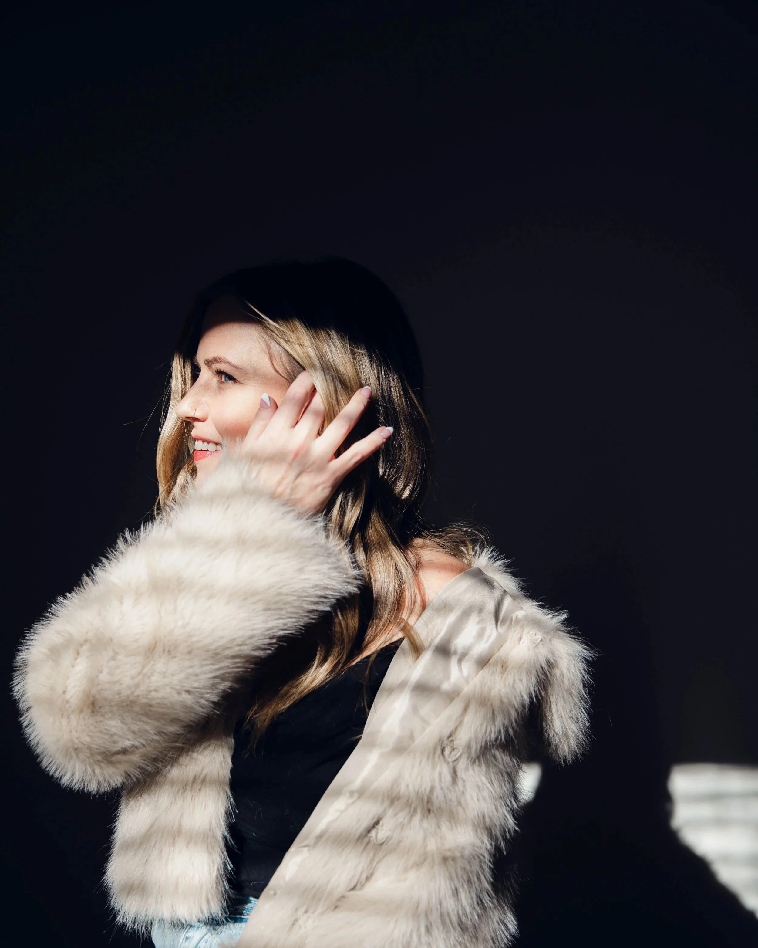 A smiling woman with wavy blonde hair wearing a faux fur coat against a dark background.