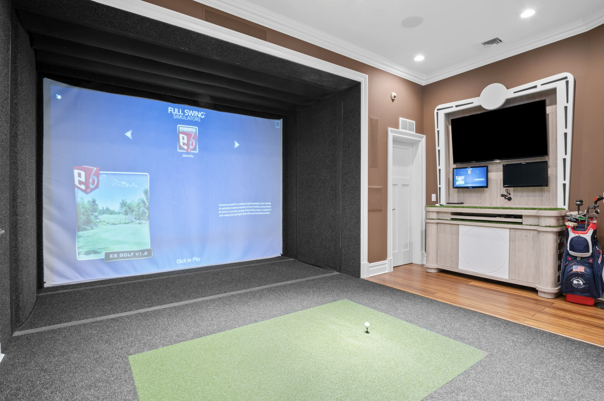 Indoor golf simulator room with a large screen showing a golf game, a small green mat with a golf ball, a wooden cabinet below a mounted large TV, and a golf bag with clubs on the right side.
