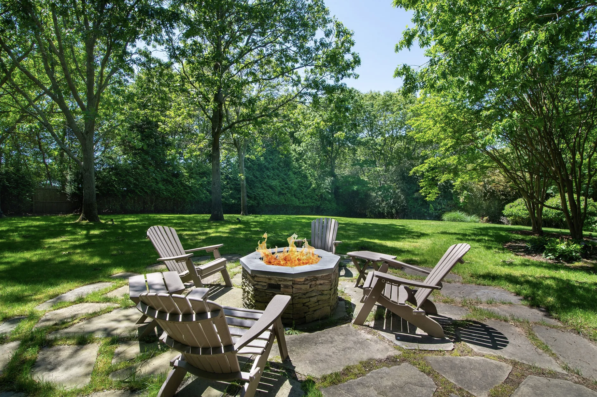 A backyard with a fire pit surrounded by six wooden Adirondack chairs on a stone patio, with green grass and trees in the background under a blue sky.