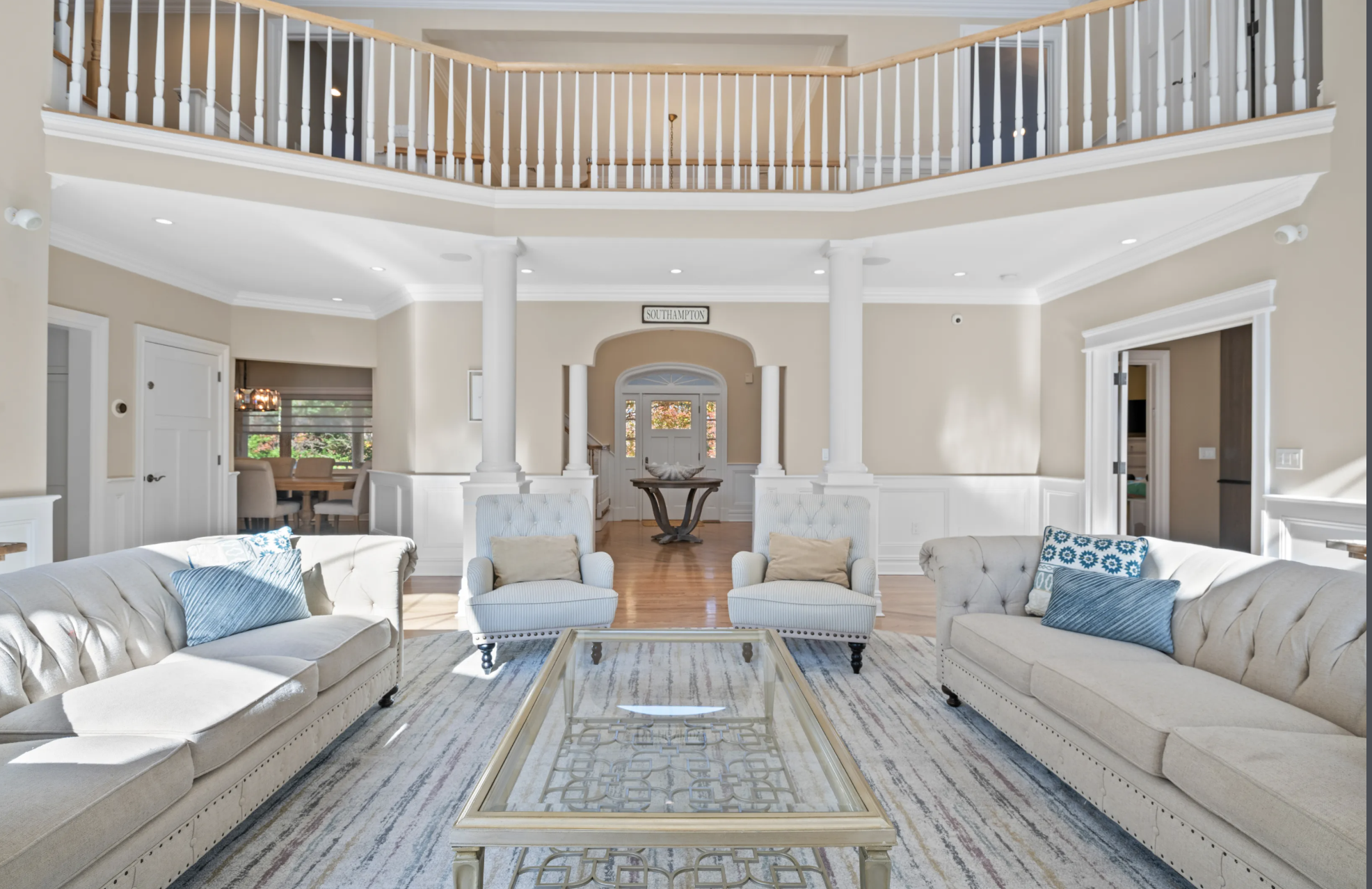 Elegant living room with two white sofas, two light blue armchairs, a glass coffee table, cream-colored walls, and a wood staircase in the background.