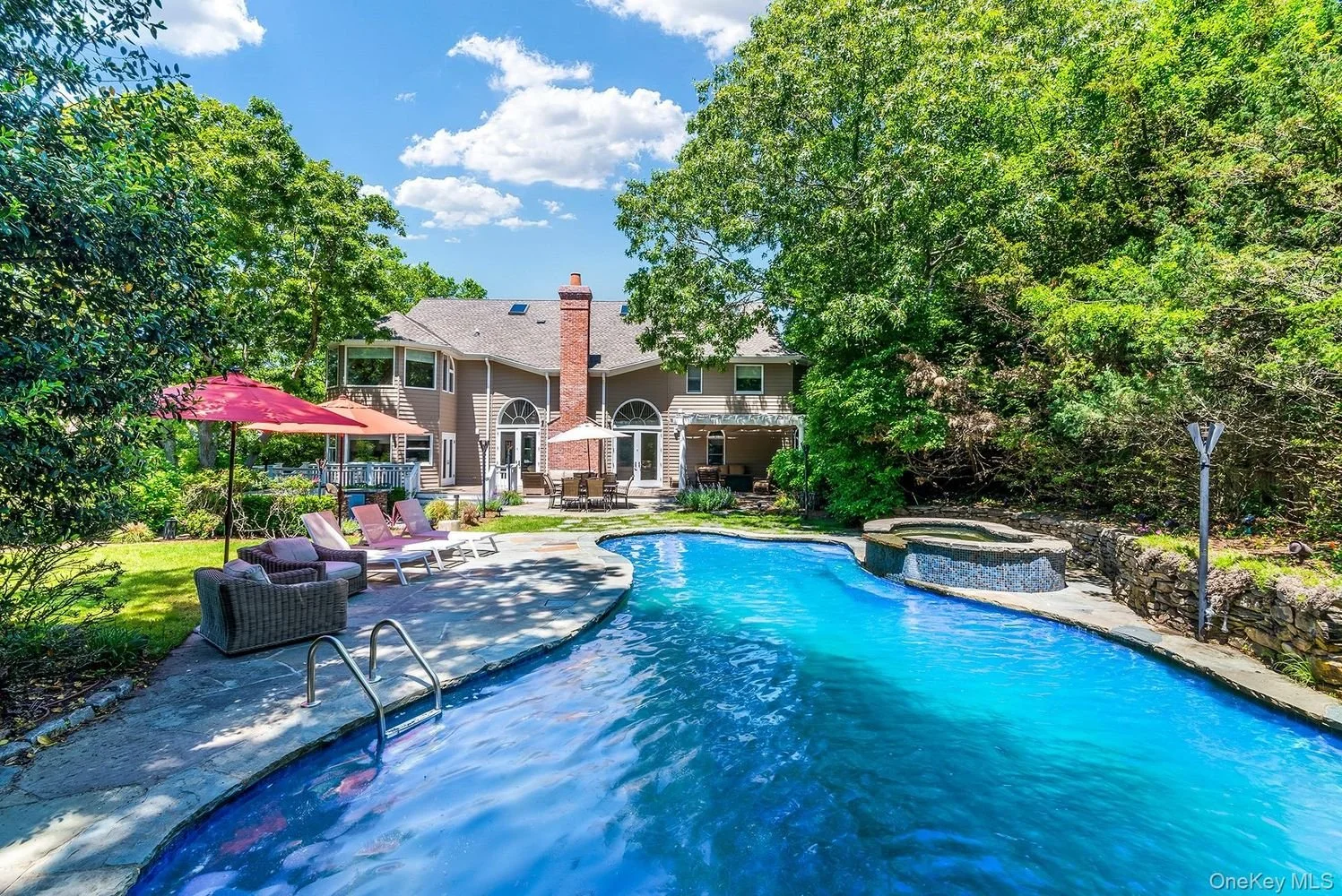 Backyard view of a house with a large swimming pool, lounge chairs, umbrellas, and surrounding greenery under a partly cloudy sky.
