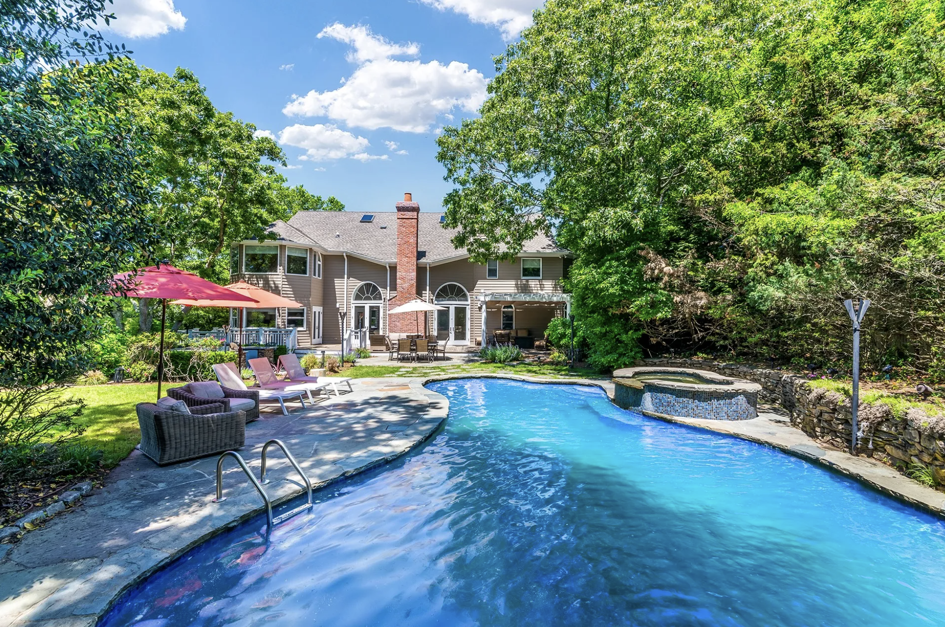 Backyard with a swimming pool, lounge chairs, umbrellas, and a two-story house with a chimney surrounded by green trees and a blue sky.