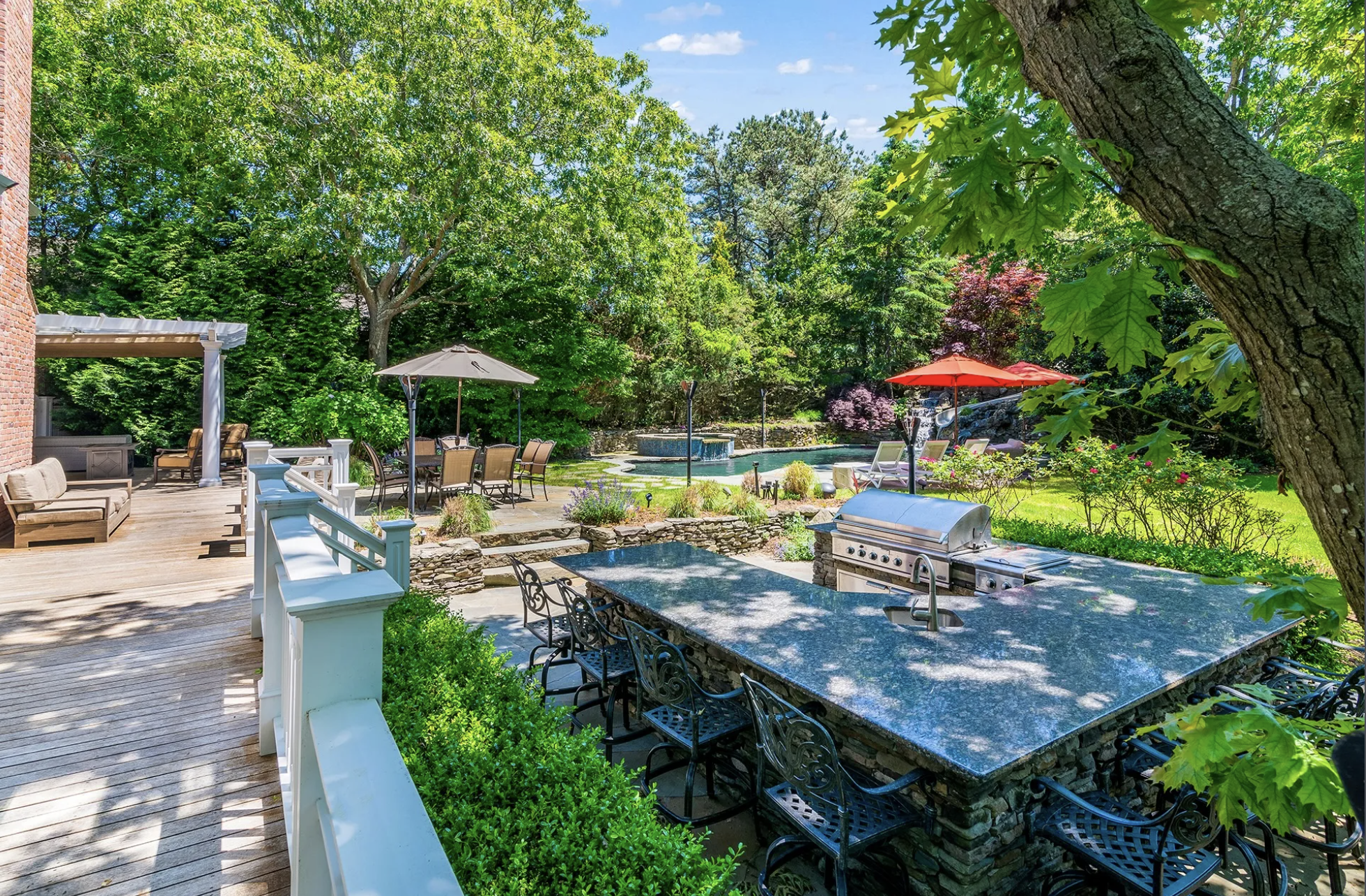 Backyard with a wooden deck, outdoor seating, a stone outdoor kitchen counter with bar stools, a swimming pool with a hot tub, patio umbrellas, and lush green trees and bushes.