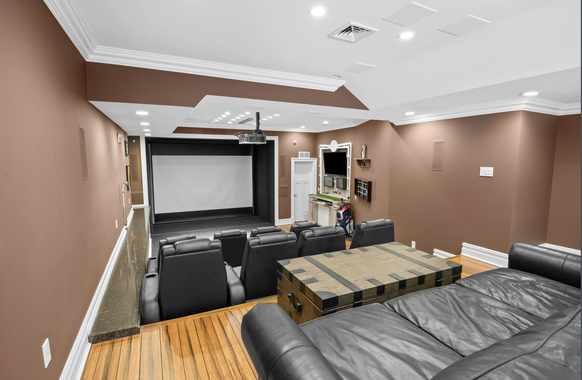 A cozy home theater room with a large projection screen, surrounded by black framing, and a ceiling-mounted projector. Brown walls with white crown molding, wooden flooring, and a mix of black leather seating including a sofa and multiple armchairs a