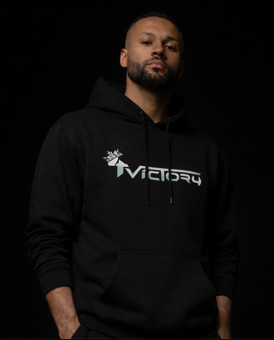 Un homme porte un sweat à capuche noir avec le logo '4Victory' sur la poitrine, pose devant un fond noir.