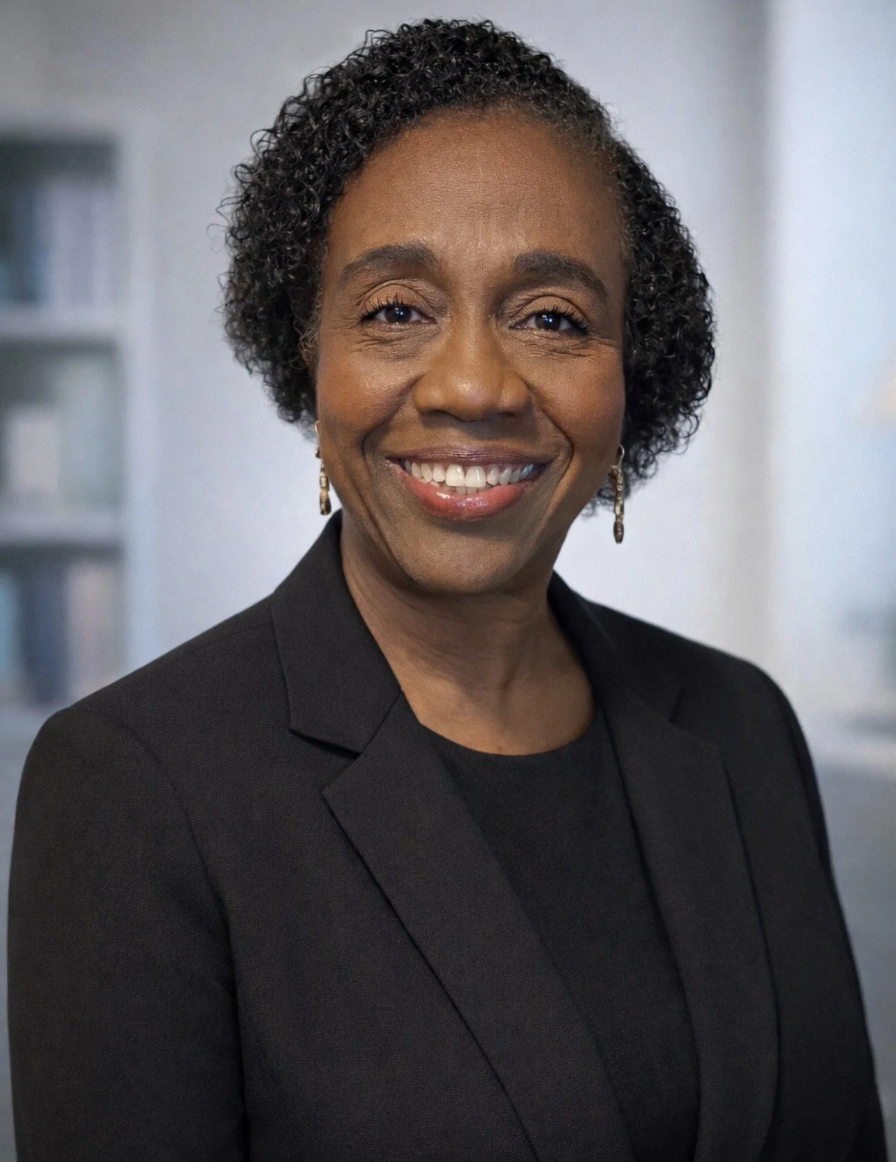 A middle-aged Asian man with gray hair sits on a bench against a plain, beige wall, looking to his left with a calm expression.