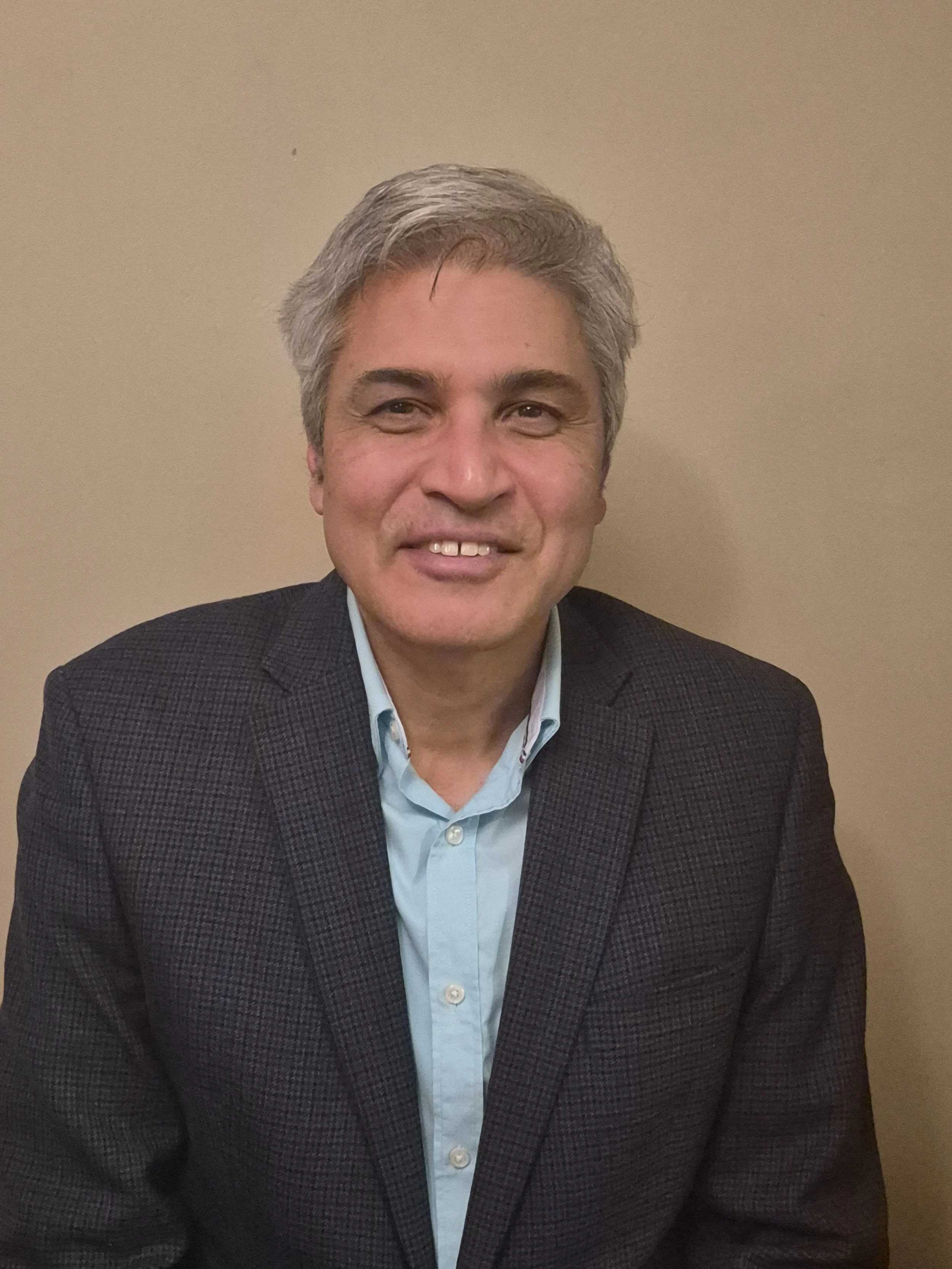 A middle-aged man with gray hair, wearing a light blue collared shirt and dark blazer, smiling and sitting against a beige background.