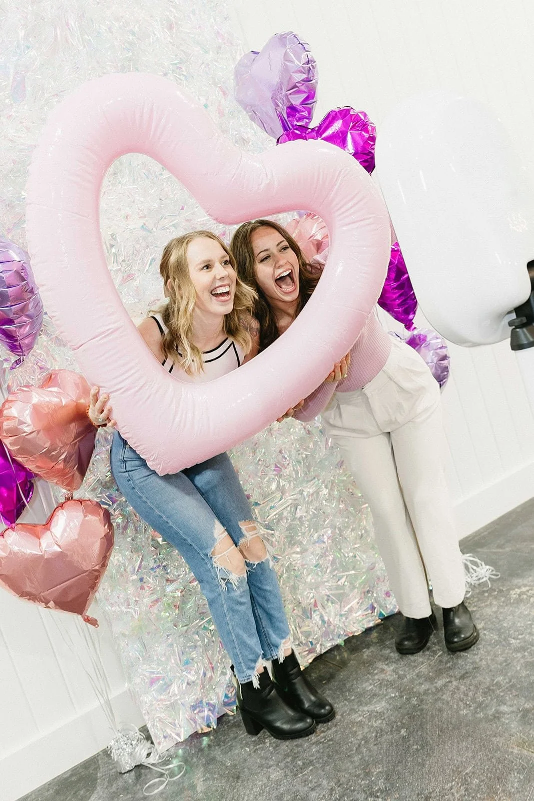Two women with happy expressions holding a large pink heart-shaped balloon, surrounded by smaller heart-shaped balloons and iridescent decorations.