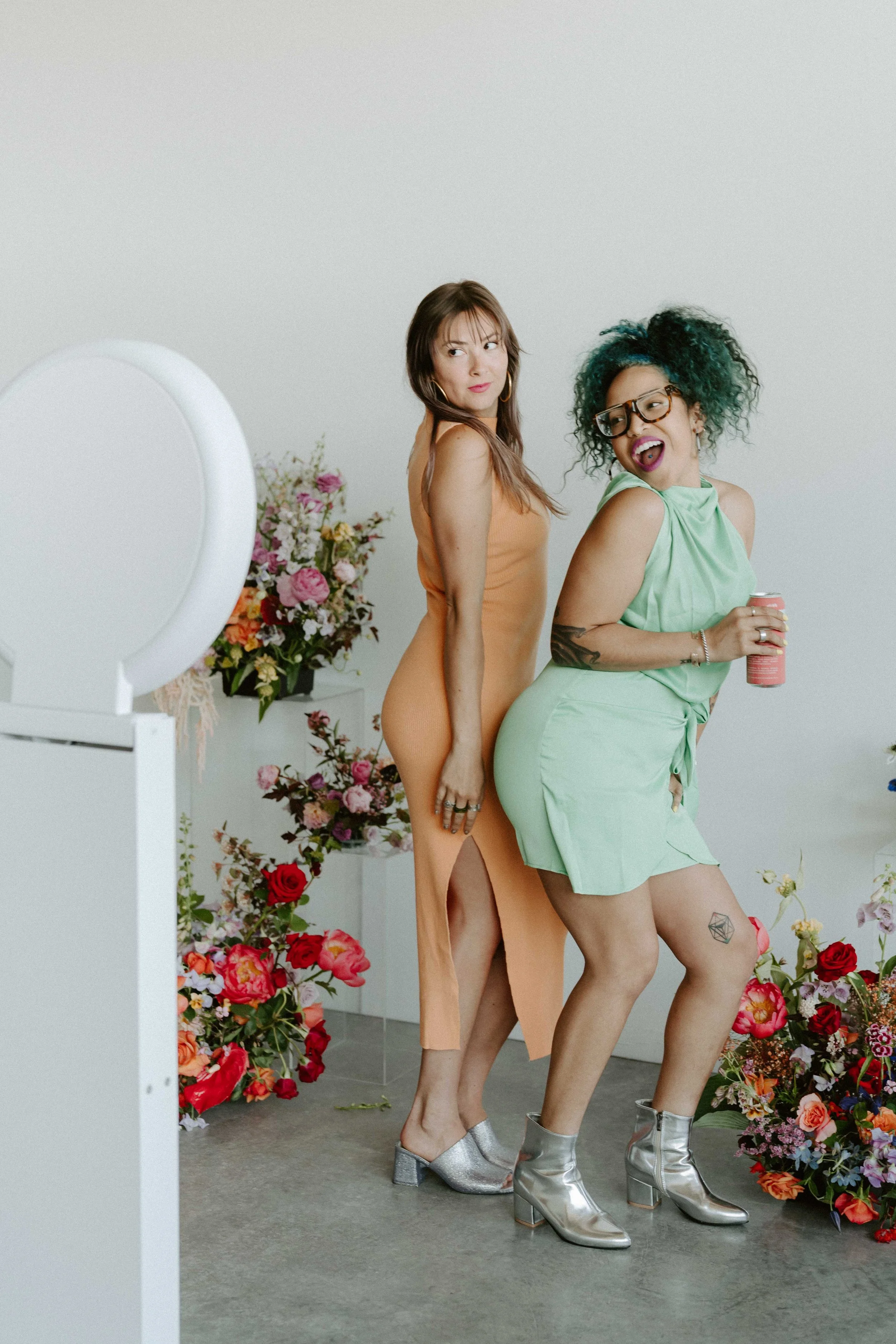 Two women standing among colorful flowers with a mirror and white wall in the background. One woman has brown hair, glasses, and wears a green dress; the other has brown hair and wears an orange dress.