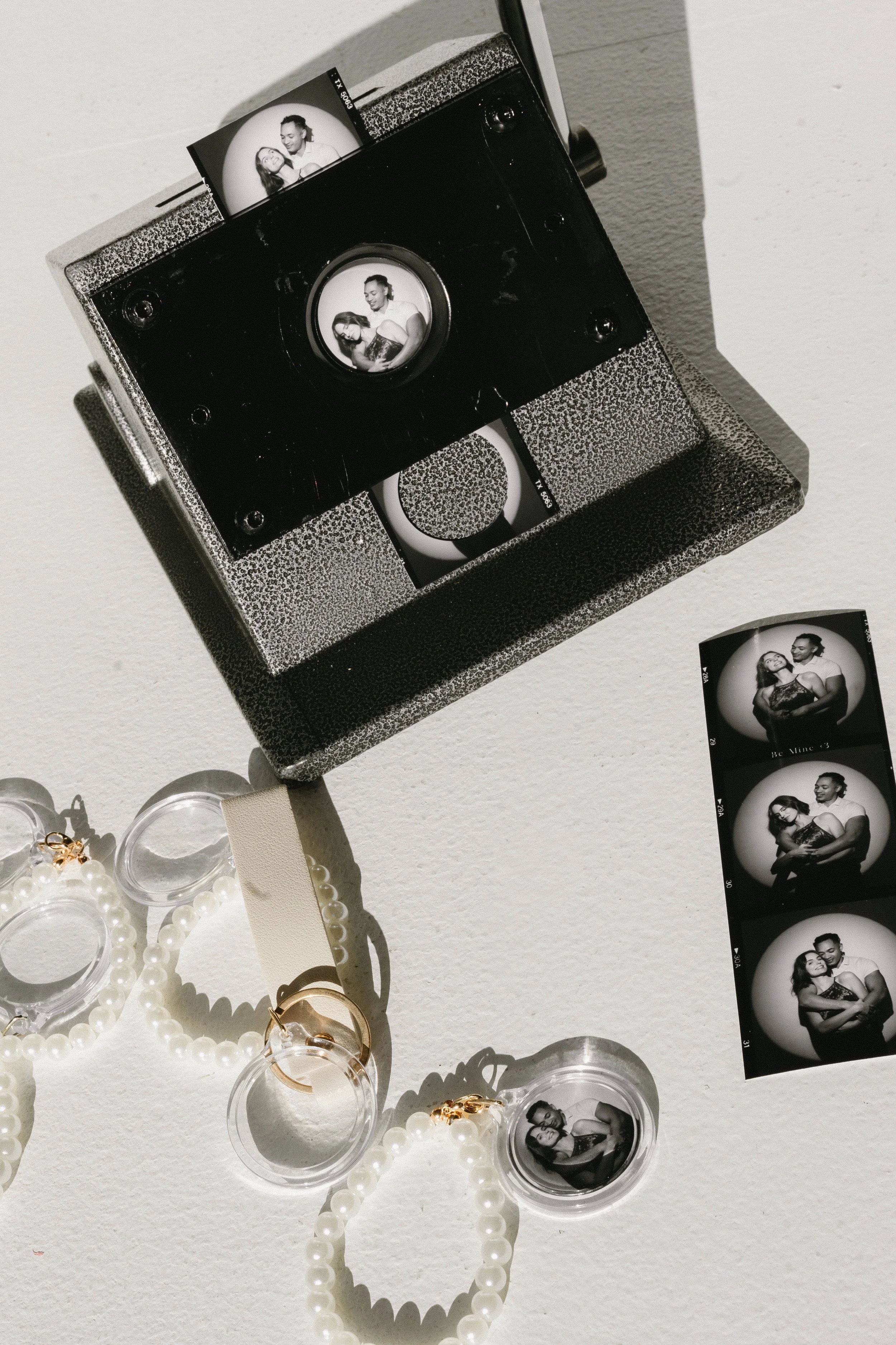 A vintage camera with black and silver textures displays black and white photo strips showing a couple's portrait. Surrounding the camera are pearl jewelry pieces, plastic containers with jewelry, and a strip of black and white contact sheets with more photos of the same couple.