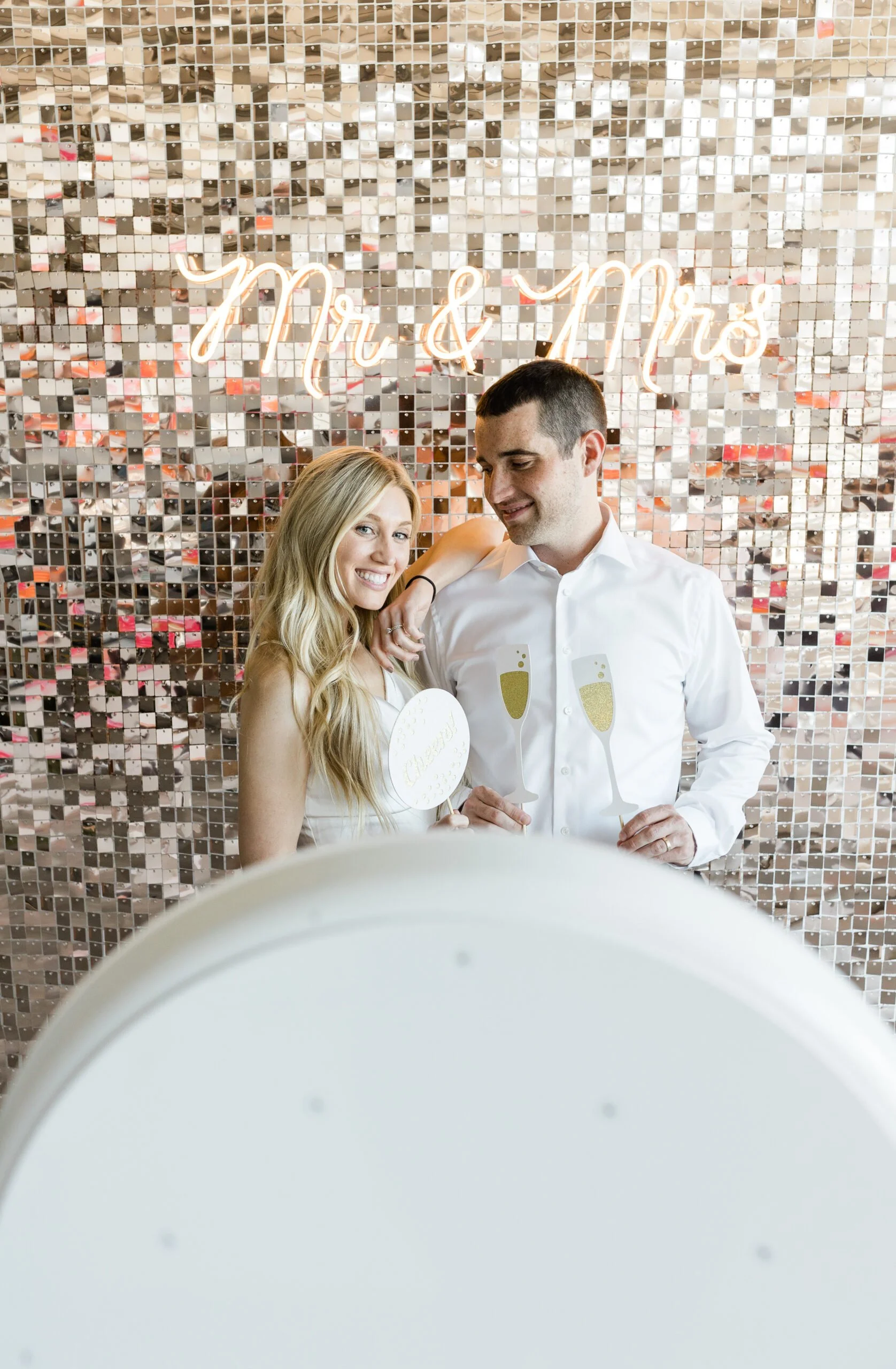 A couple at a celebration, with a sparkling, reflective mosaic wall and a neon sign that reads "Mr & Mrs" behind them. The woman has long blond hair and is smiling while holding a white heart-shaped sign. The man has short dark hair and is wearing a white shirt, holding a champagne flute cutout feature.