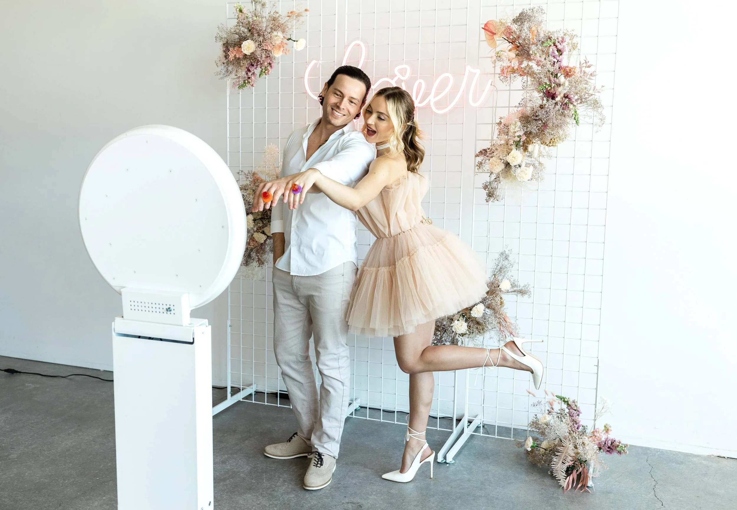 A couple taking a selfie in front of a floral backdrop with a neon sign that says "Cher," at a celebration or event. The woman is wearing a nude-colored tulle dress and white high heels, while the man is wearing a white shirt and beige pants.