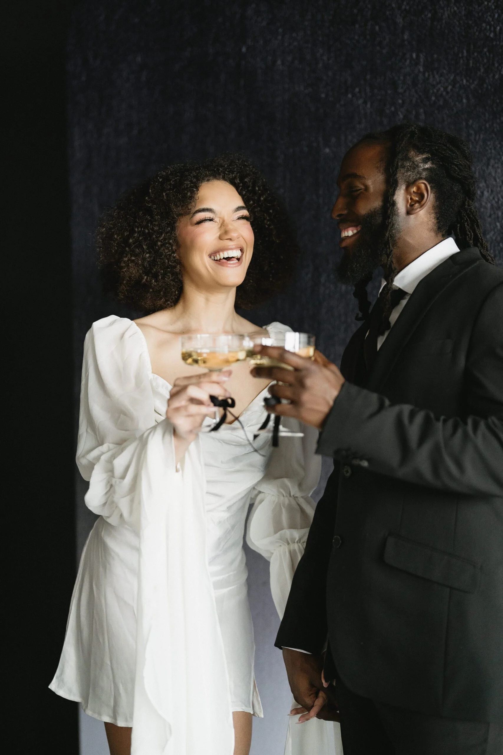 A woman and a man are smiling and holding glasses of champagne. The woman has curly hair and is wearing a white dress. The man has dreadlocks and is dressed in a black suit. They are standing against a dark textured wall.