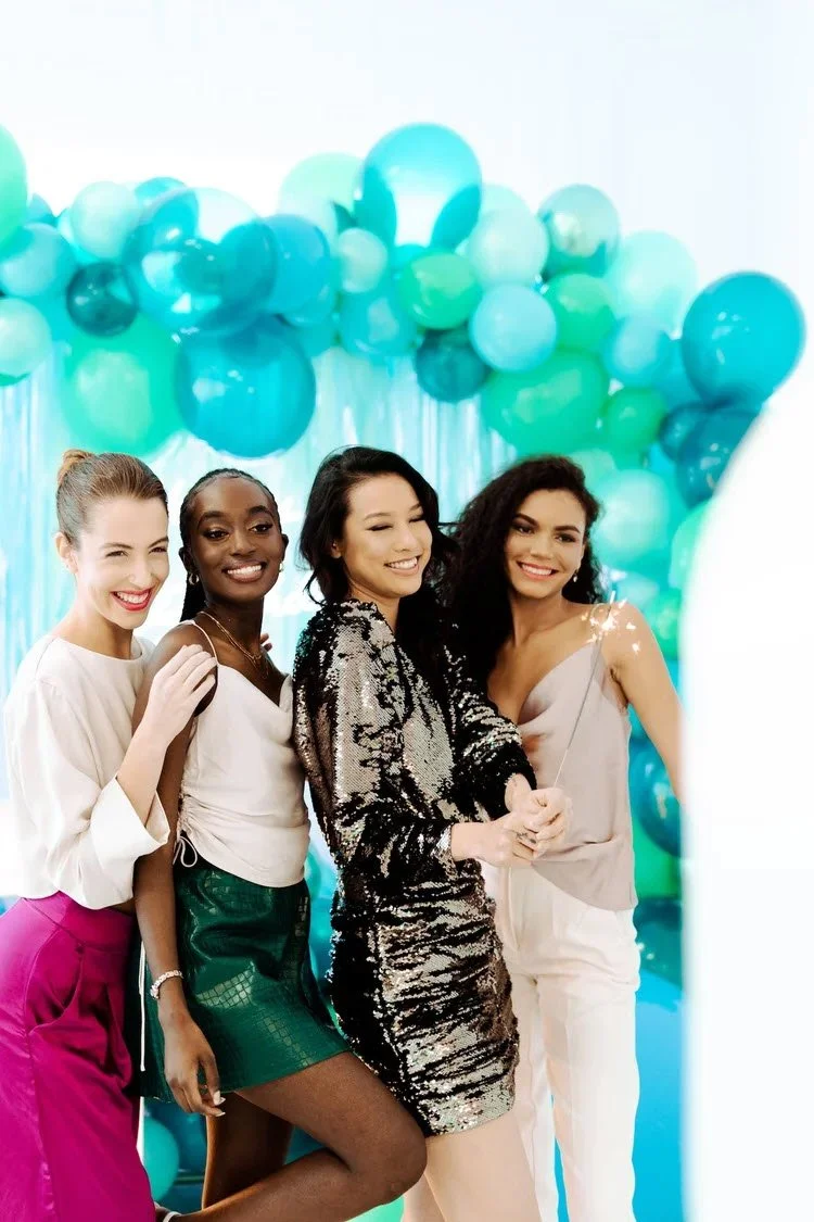 Four women smiling at a party with teal and white balloons in the background, dressed in stylish outfits.