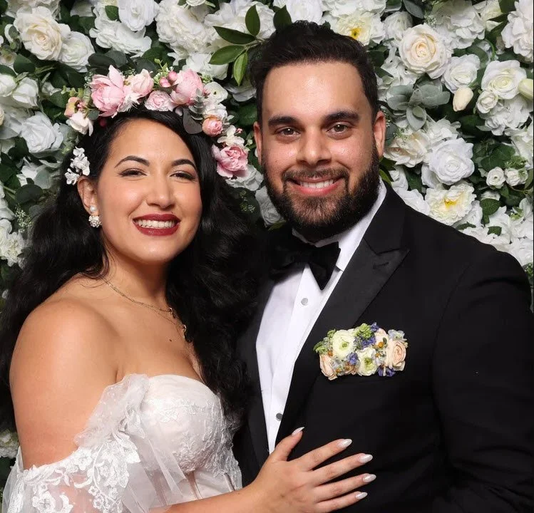 A newlywed couple dressed in wedding attire, standing in front of a floral wall, smiling at the camera. The bride is wearing a white wedding dress with lace details and a flower crown, while the groom is in a black tuxedo with a bow tie and boutonniere.