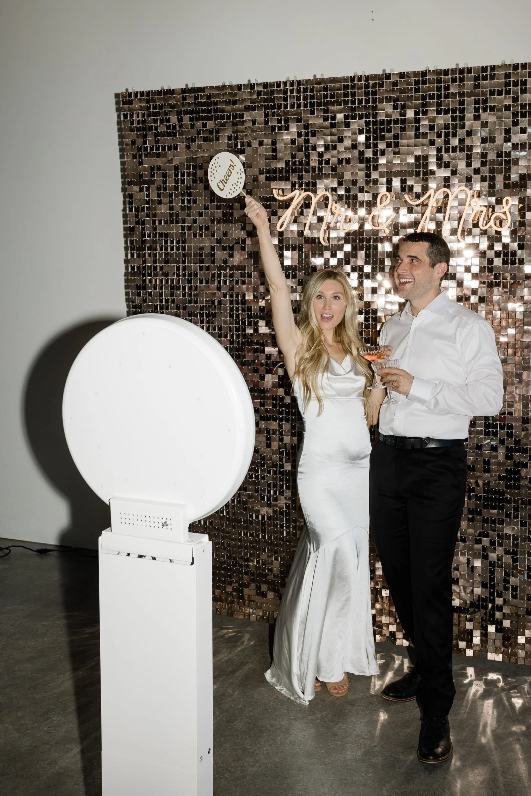 A couple dressed in wedding attire standing in front of a decorative backdrop, with a photo booth prop and an open photo booth in the foreground.