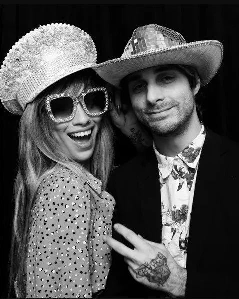 Two people at a party wearing sparkling hats and glasses, posing against a dark background.