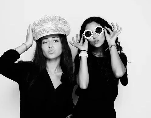 Two young women posing with playful accessories; one wearing a bejeweled hat and the other holding flower-shaped sunglasses.