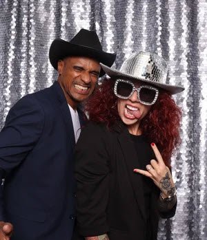 Two people posing in front of a silver sequin backdrop, wearing fun accessories and making playful expressions.