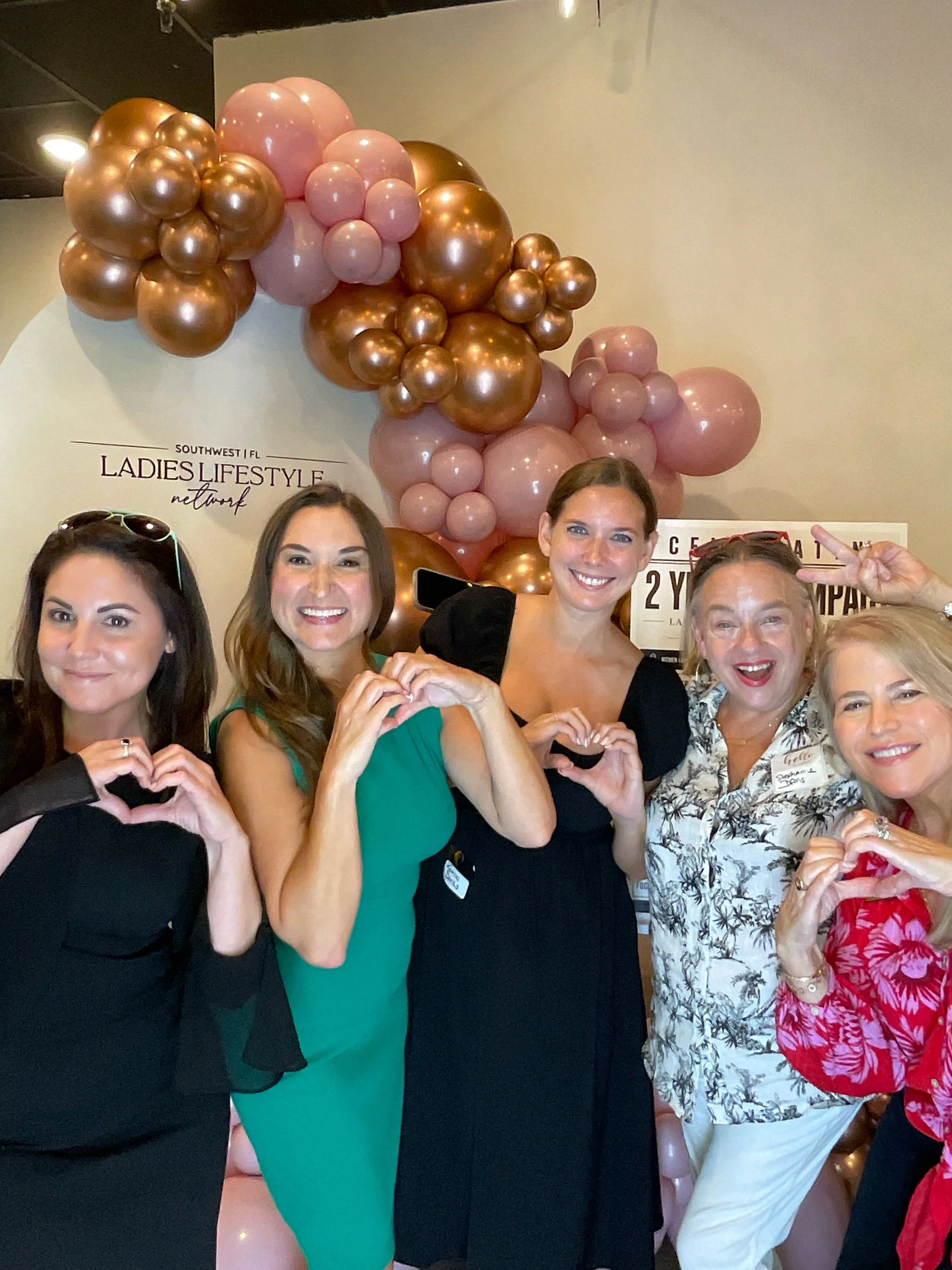A group of women smiling and posing for a photo, with balloons in gold and pink colors in the background, at an event celebrating a Ladies Lifestyle Network. They are making heart signs with their hands.