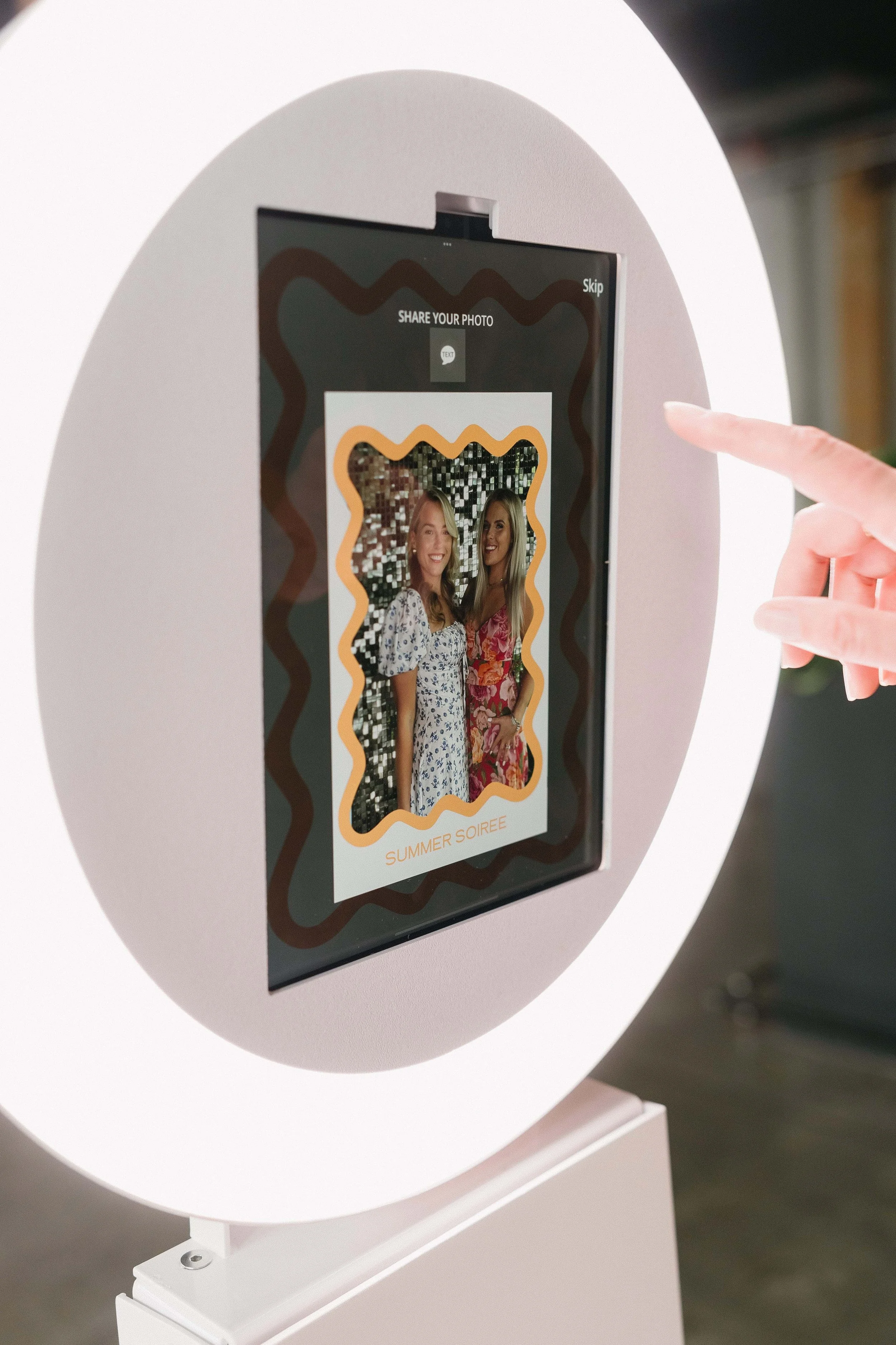 A photo booth with a large circular white frame and a screen that shows a photo of two smiling women in colorful dresses. The screen has decorative borders around the photo with the text 'Summer Soiree' at the bottom. A person's hand is pointing towards the screen.